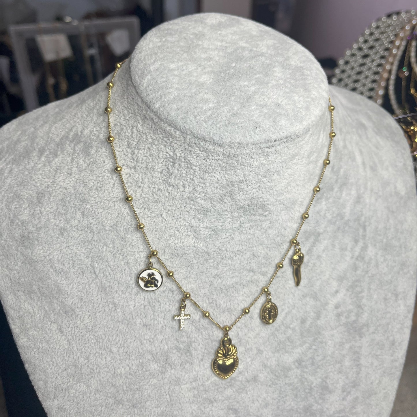 Collana Zoe Charms con Angelo in acciaio premium placcato oro con ciondoli multipli e charm angelo, indossata su busto espositore grigio