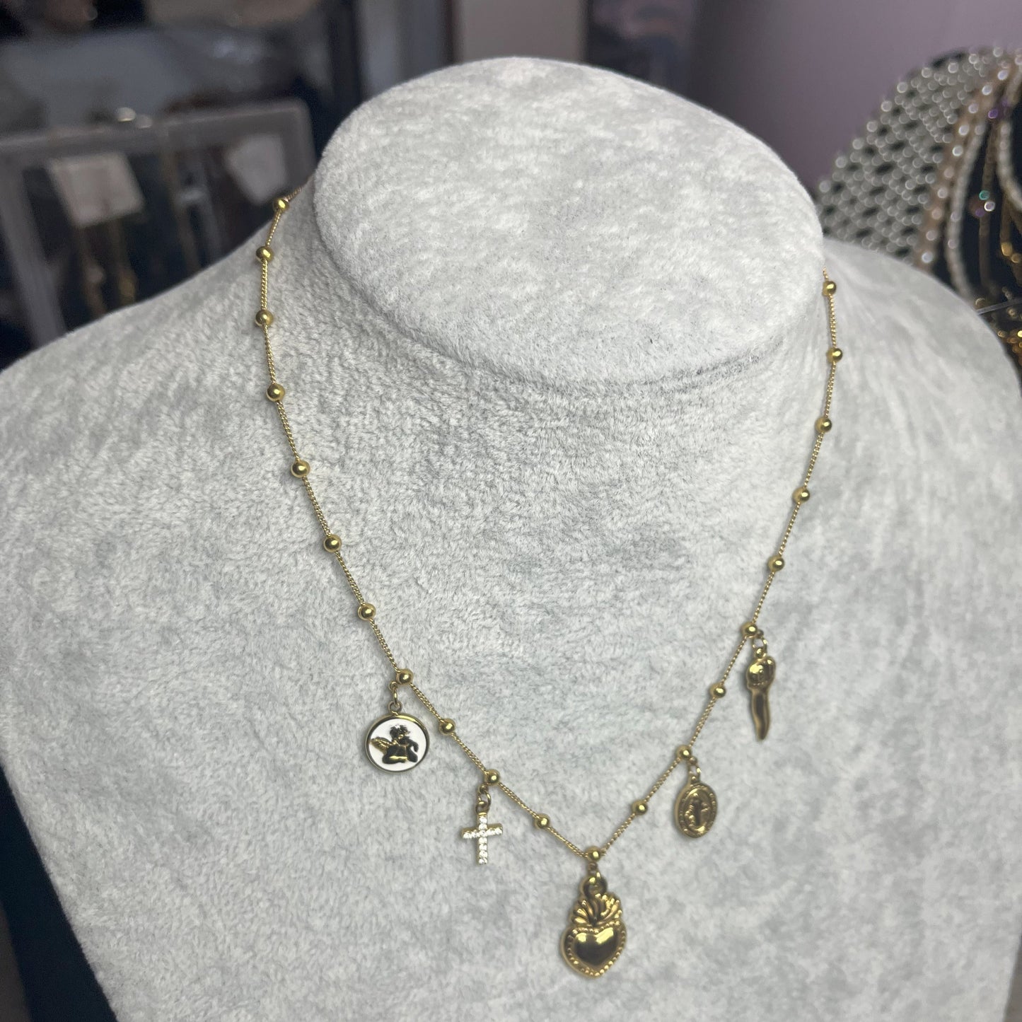 Collana Zoe Charms con Angelo in acciaio premium placcato oro con ciondoli multipli e charm angelo, indossata su busto espositore grigio