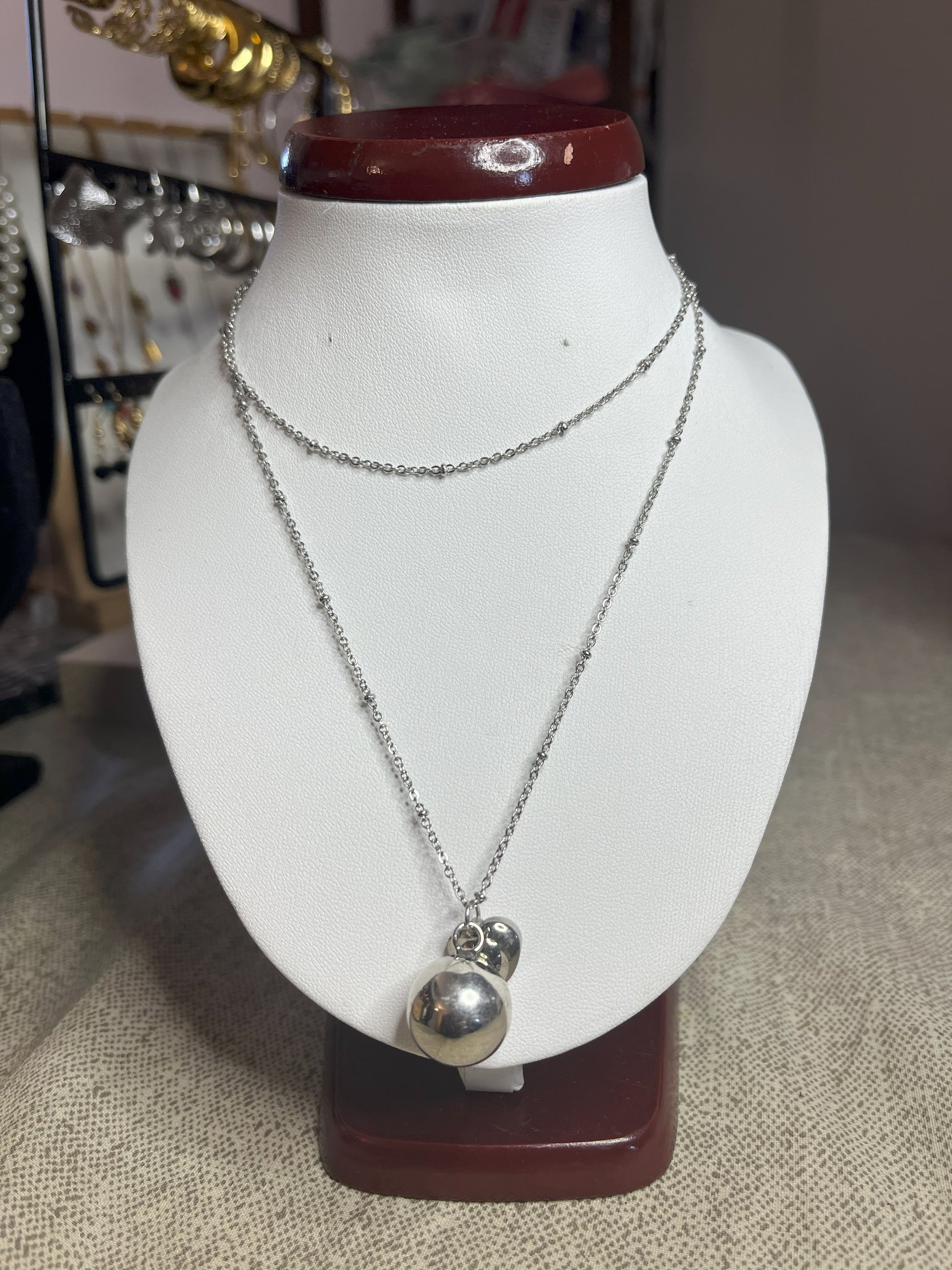 Collana Doppio Giro con sfera a sonaglio e ciondolo cuore in acciaio premium, disponibile nelle variante placcato argento, esposta su busto espositore bianco