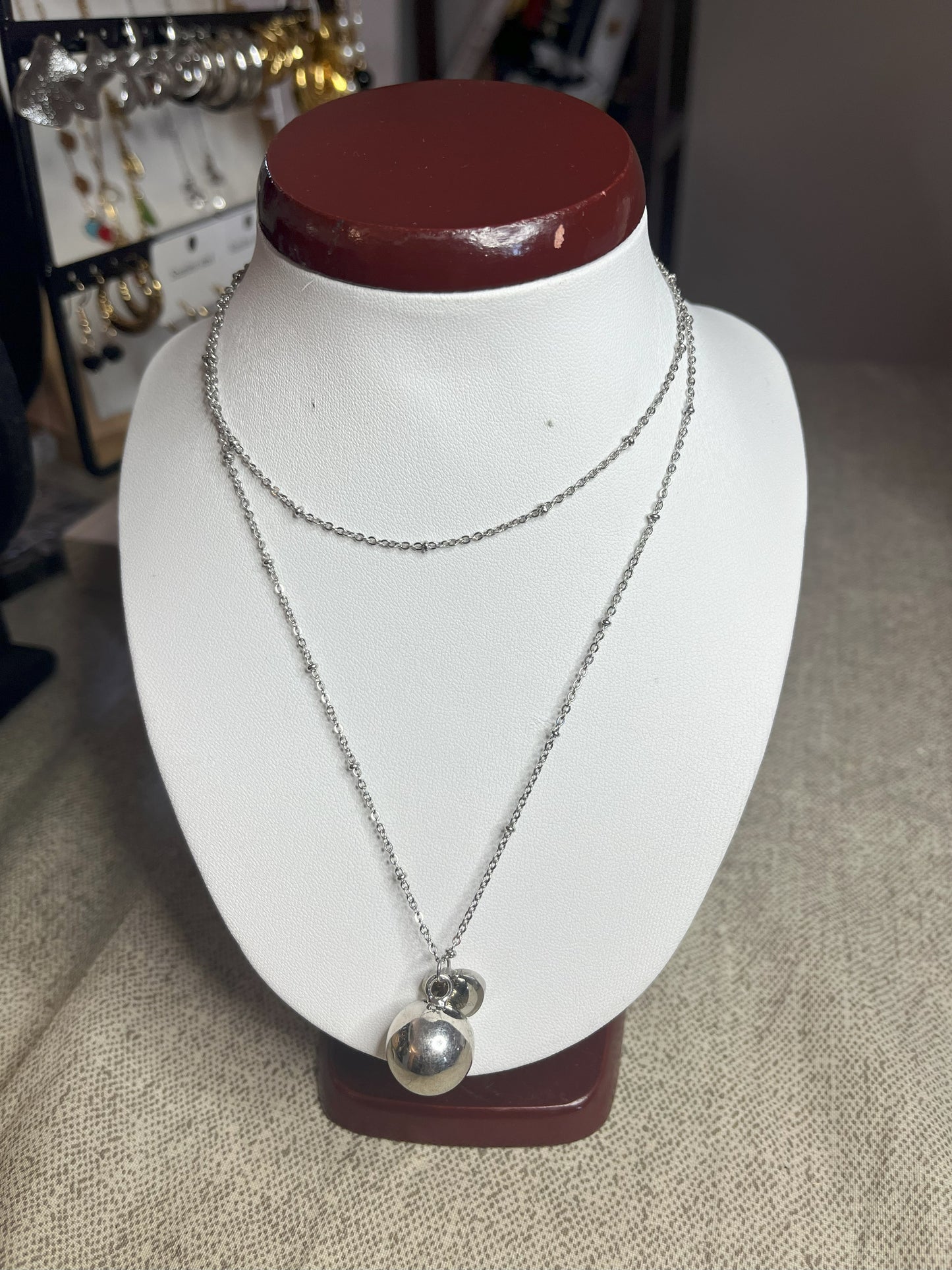 Collana Doppio Giro con sfera a sonaglio e ciondolo cuore in acciaio premium, disponibile nelle variante placcato argento, esposta su busto espositore bianco
