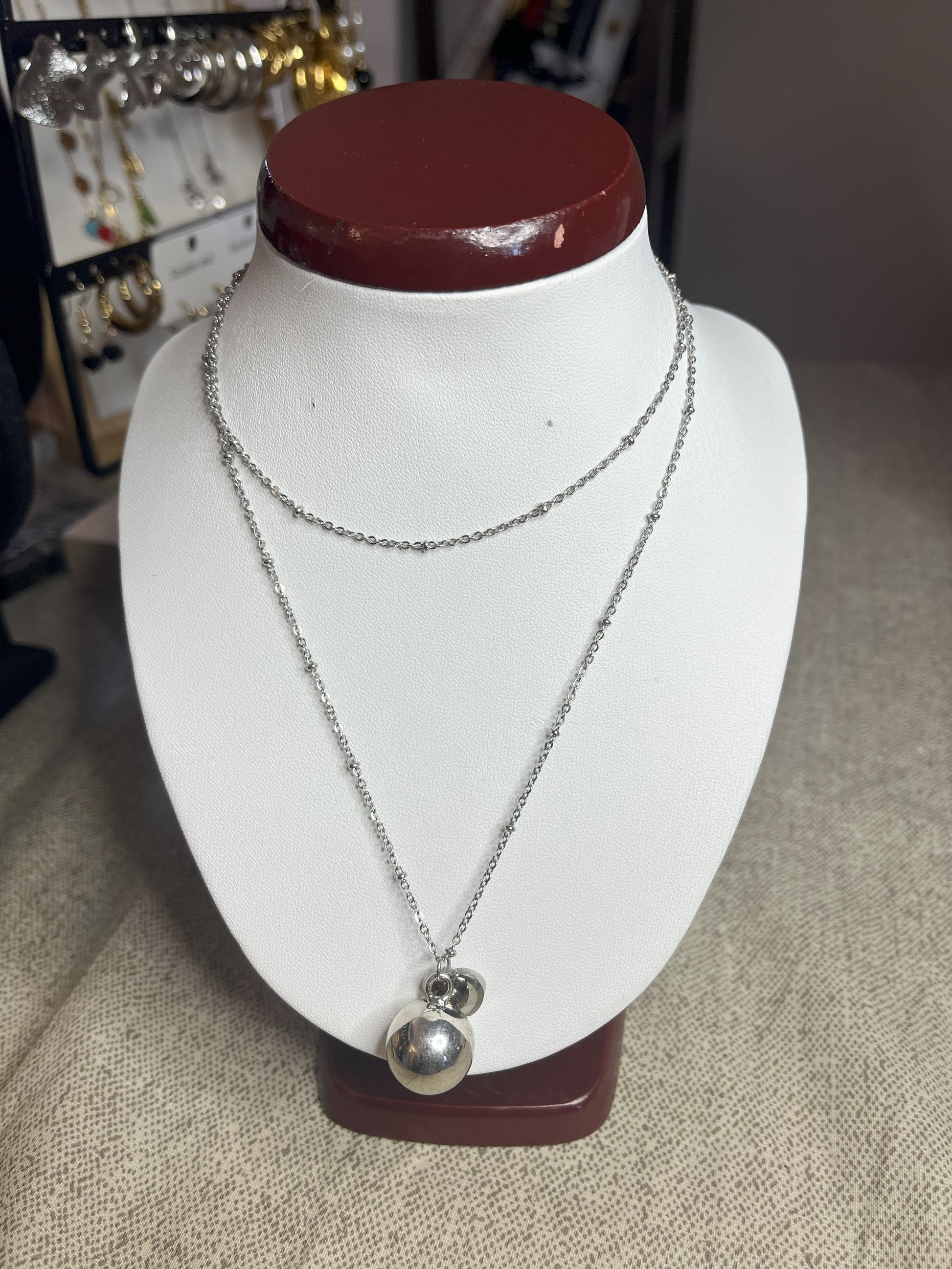 Collana Doppio Giro con sfera a sonaglio e ciondolo cuore in acciaio premium, disponibile nelle variante placcato argento, esposta su busto espositore bianco