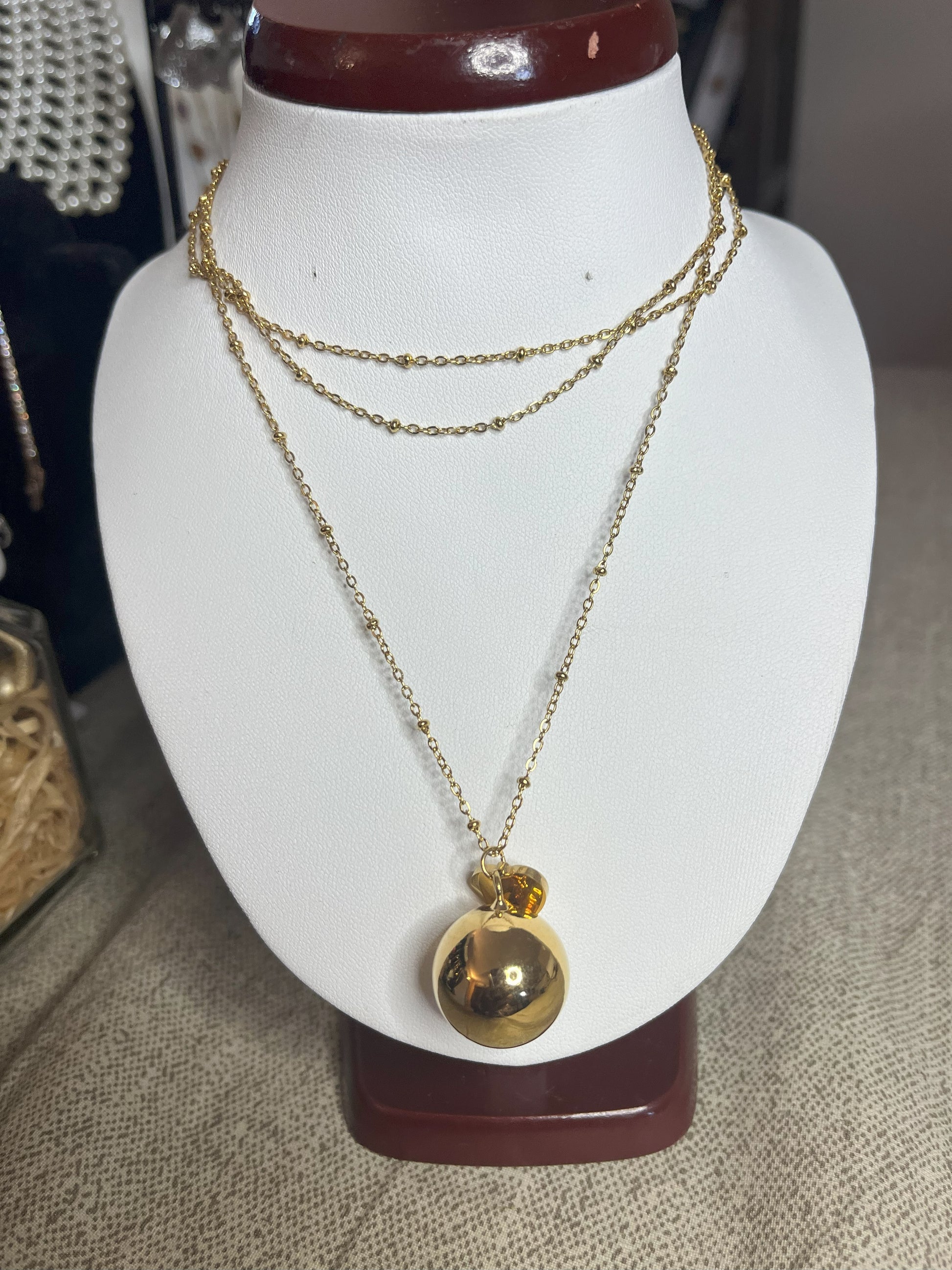 Collana Doppio Giro con sfera a sonaglio e ciondolo cuore in acciaio premium, disponibile nelle variante placcato oro, esposta su busto espositore bianco