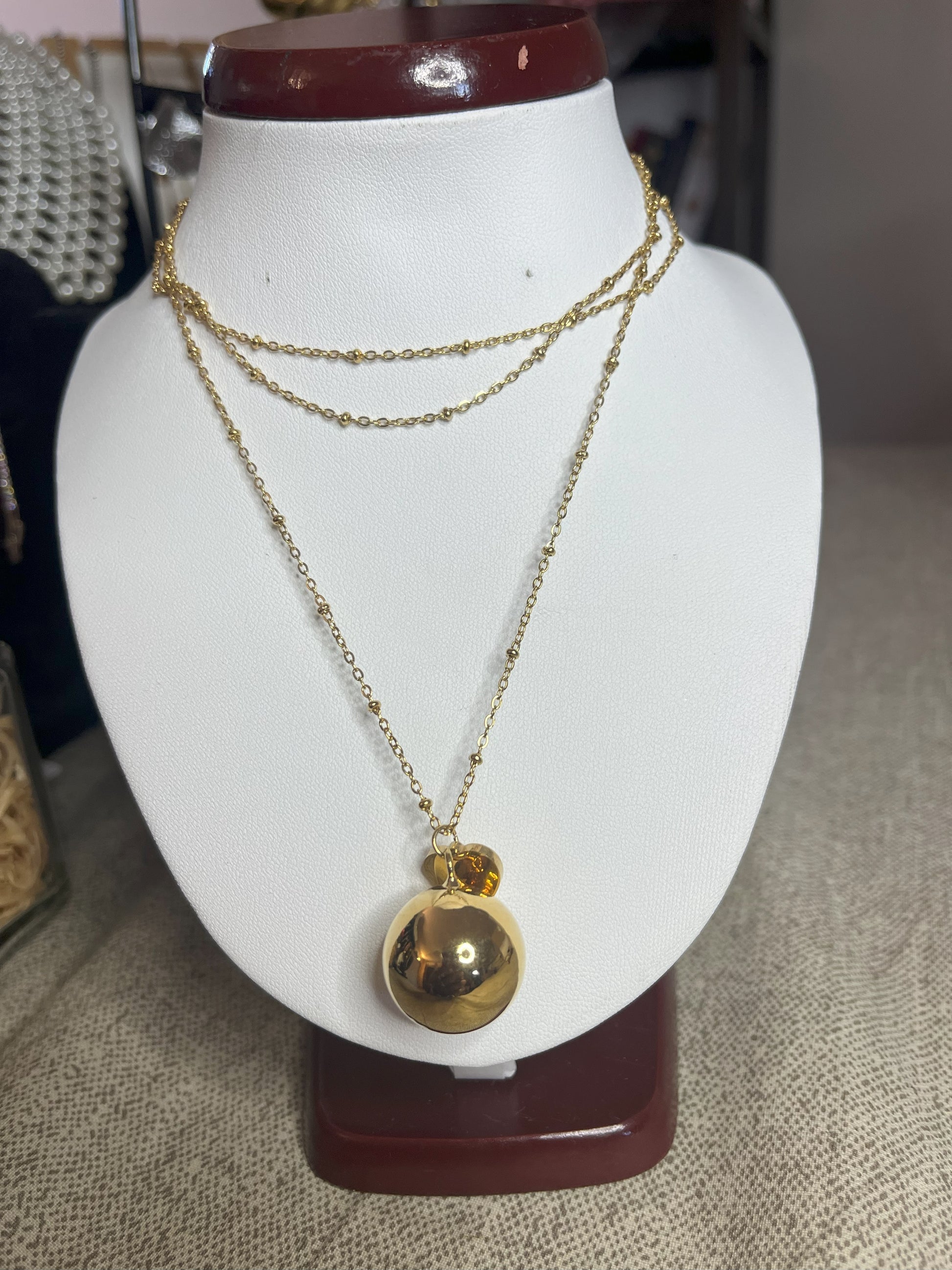 Collana Doppio Giro con sfera a sonaglio e ciondolo cuore in acciaio premium, disponibile nelle variante placcato oro, esposta su busto espositore bianco