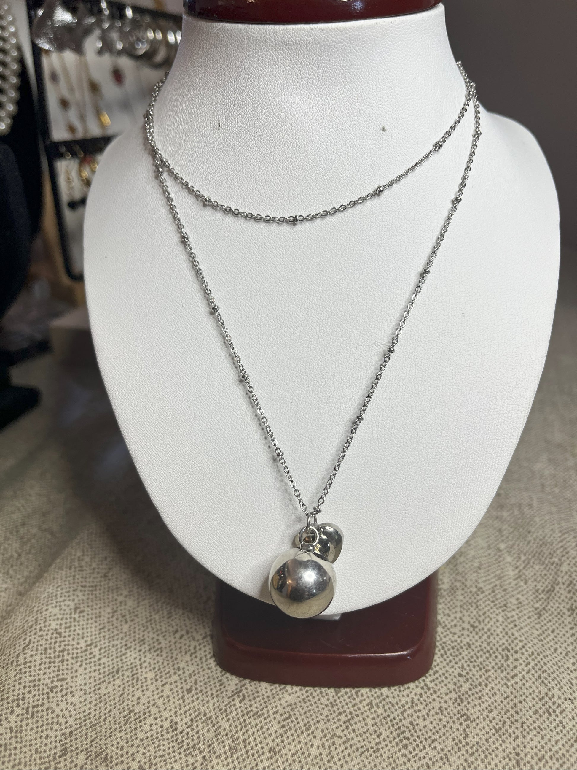 Collana Doppio Giro con sfera a sonaglio e ciondolo cuore in acciaio premium, disponibile nelle variante placcato argento, esposta su busto espositore bianco
