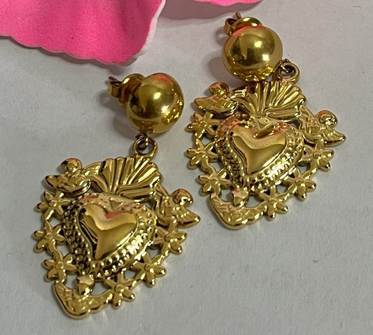 Orecchini dorati a forma di sacro cuore con dettagli intarsiati e lavorazione artigianale su sfondo con fiore rosa