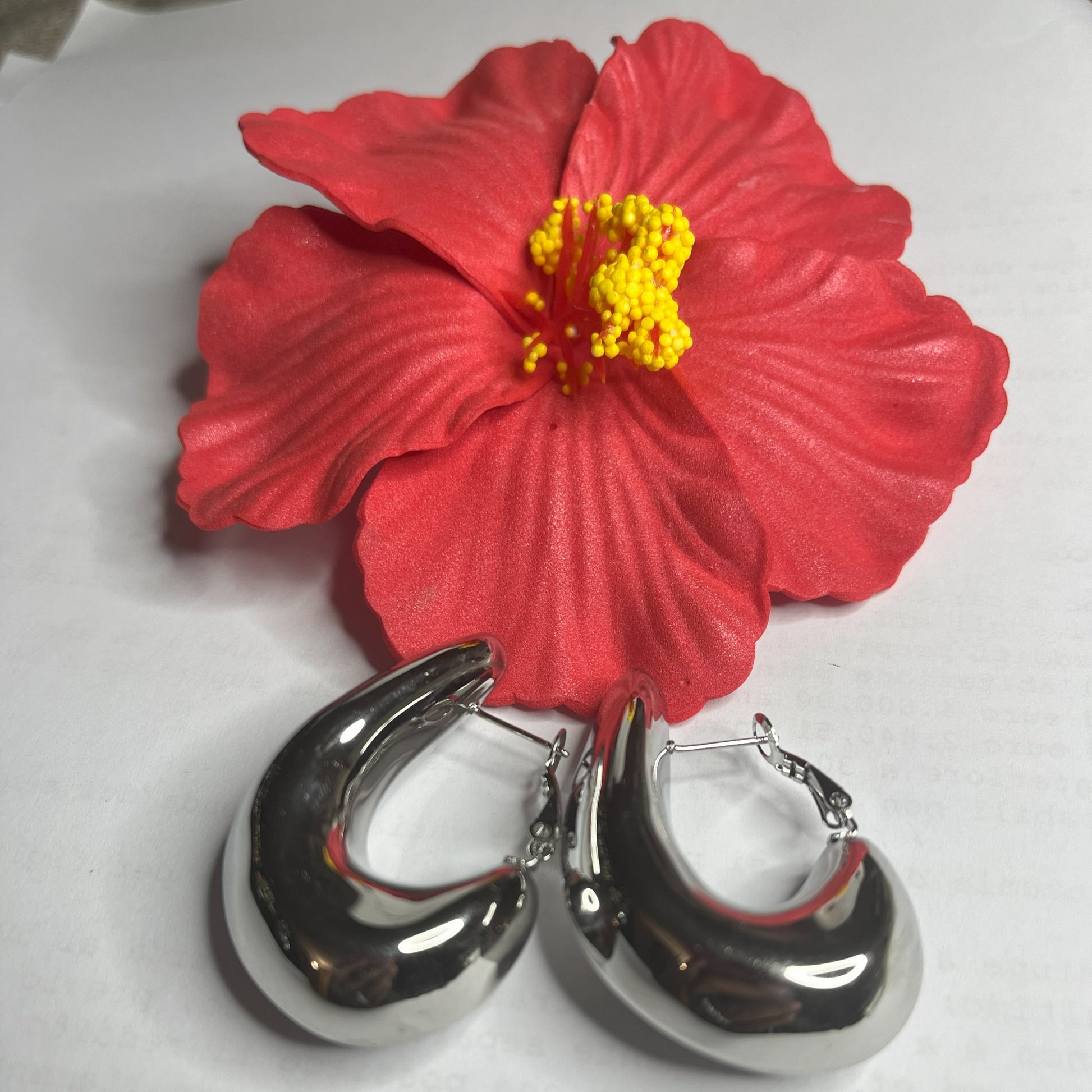 Orecchini Della Schiava a cerchio in acciaio premium placcato argento con superficie liscia e brillante, chiusura a scatto sicura, fotografati su superficie bianca e fiore rosso