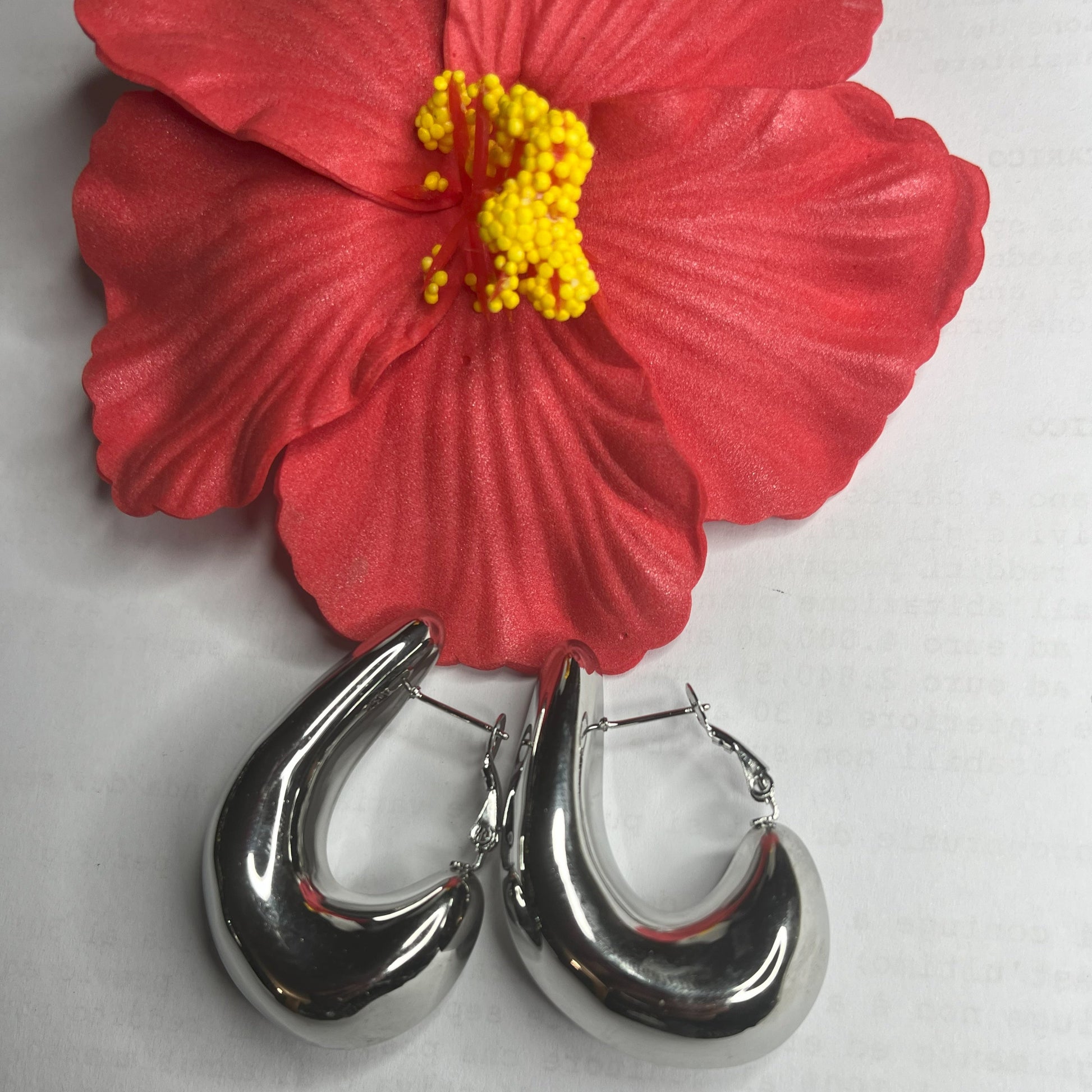 Orecchini Della Schiava a cerchio in acciaio premium placcato argento con superficie liscia e brillante, chiusura a scatto sicura, fotografati su superficie bianca e fiore rosso
