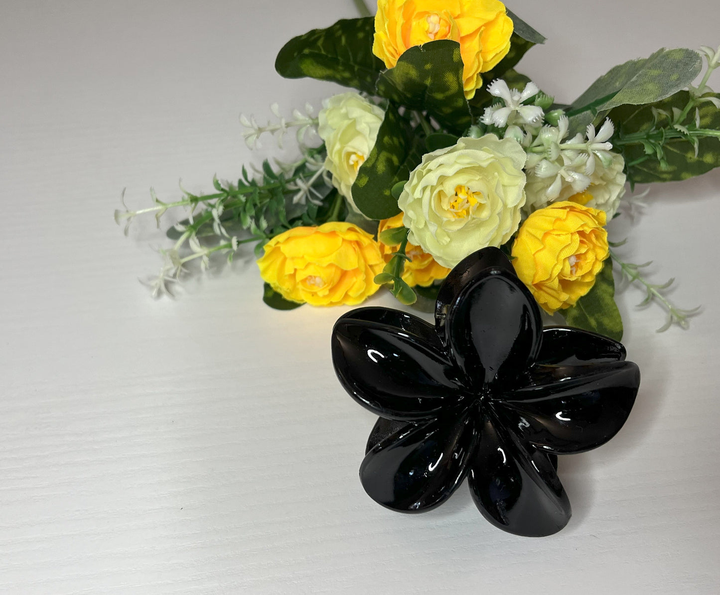 Elegante pinza hawaiana per capelli a forma di fiore con petali lucidi, posizionata accanto a un bouquet di fiori freschi gialli e bianchi