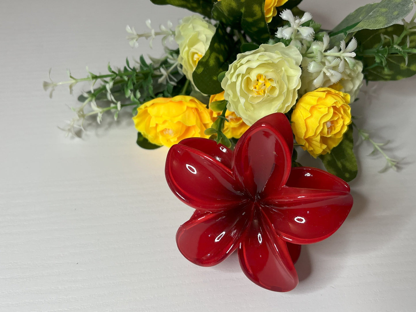 Elegante pinza hawaiana per capelli a forma di fiore con petali lucidi, posizionata accanto a un bouquet di fiori freschi gialli e bianchi