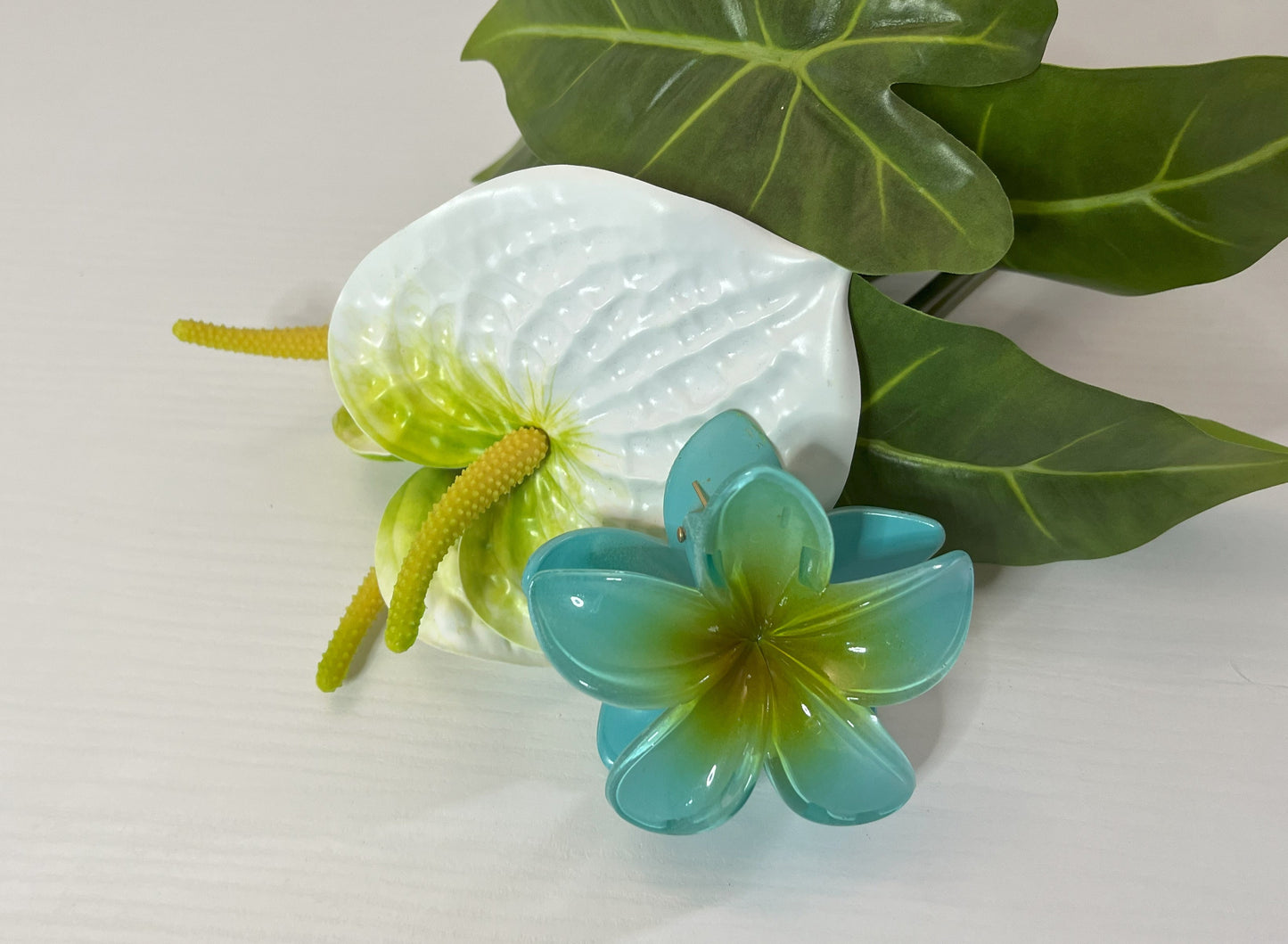 Elegante pinza hawaiana per capelli a forma di fiore con petali lucidi, posizionata accanto a un bouquet di fiori freschi gialli e bianchi