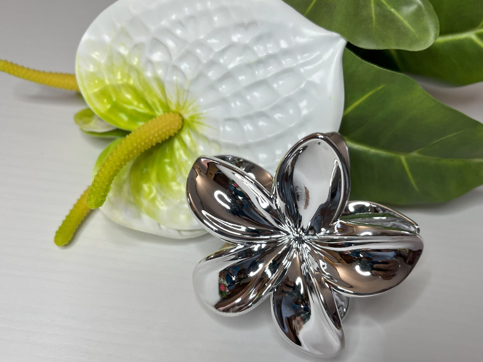 Elegante pinza hawaiana per capelli a forma di fiore con petali lucidi, posizionata accanto a un bouquet di fiori freschi gialli e bianchi