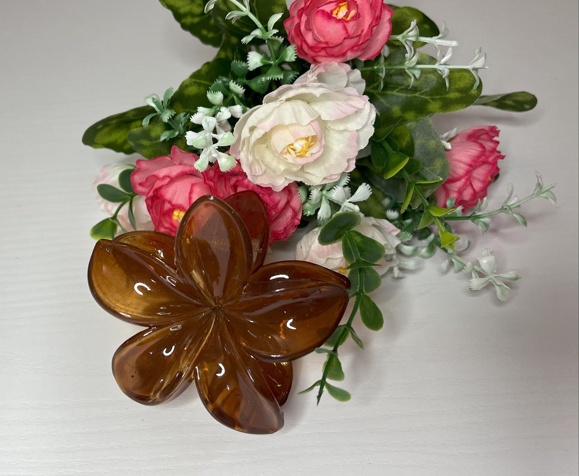 Elegante pinza hawaiana per capelli a forma di fiore con petali lucidi, posizionata accanto a un bouquet di fiori freschi gialli e bianchi