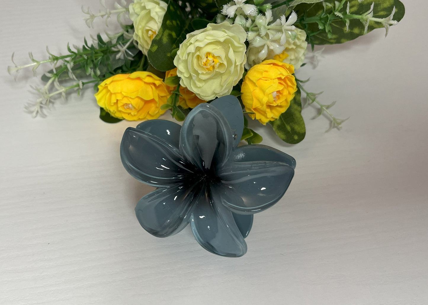 Elegante pinza hawaiana per capelli a forma di fiore con petali lucidi, posizionata accanto a un bouquet di fiori freschi gialli e bianchi