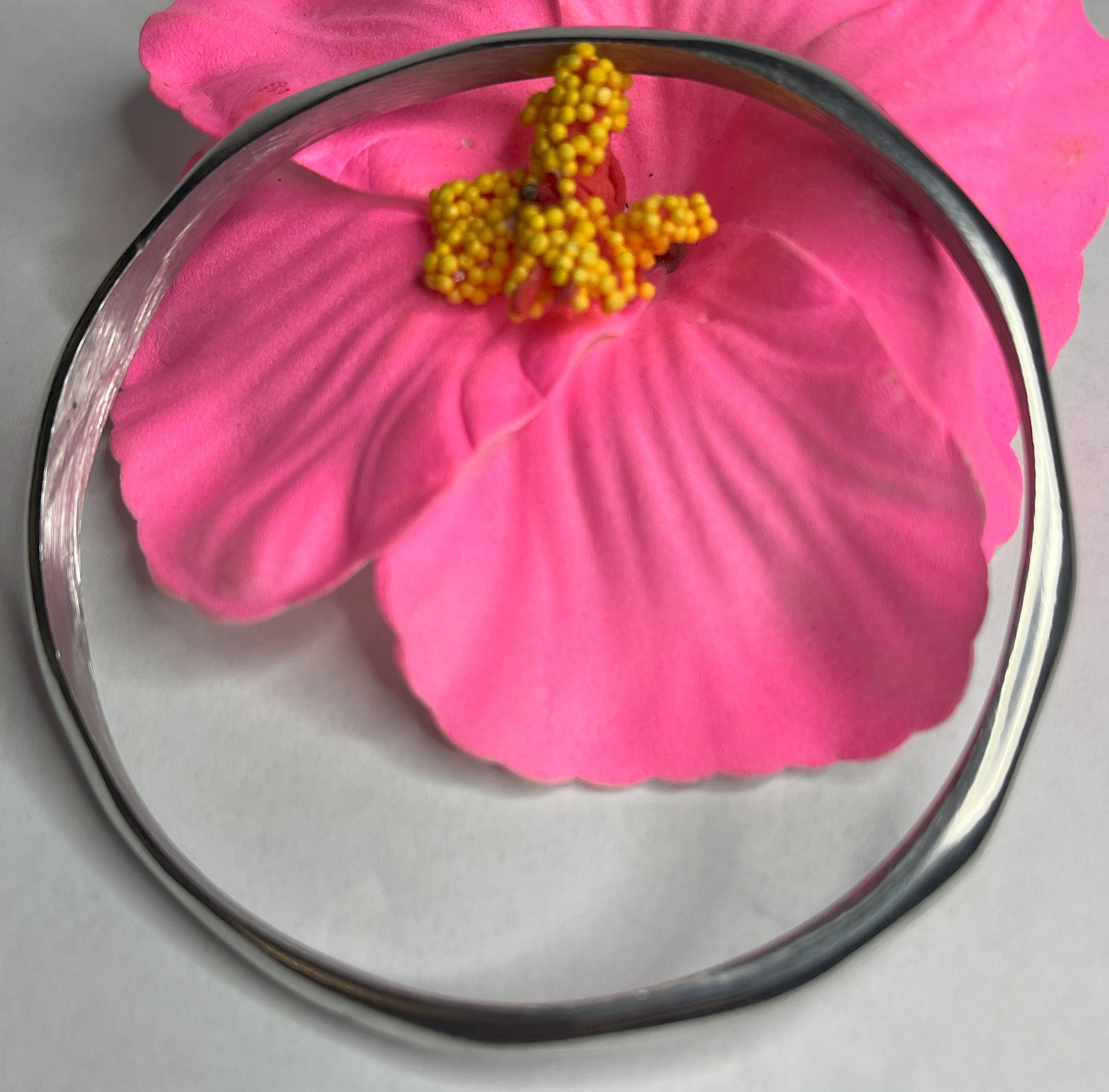 Braccialetto Rinascita in alluminio riciclato fatto a mano, design circolare minimalista con finitura lucida premium, fotografato accanto a un fiore rosa su sfondo neutro