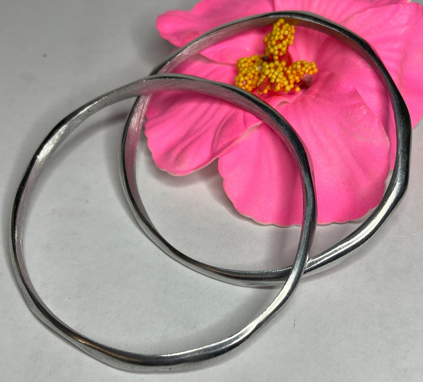 Braccialetto Rinascita in alluminio riciclato fatto a mano, design circolare minimalista con finitura lucida premium, fotografato accanto a un fiore rosa su sfondo neutro