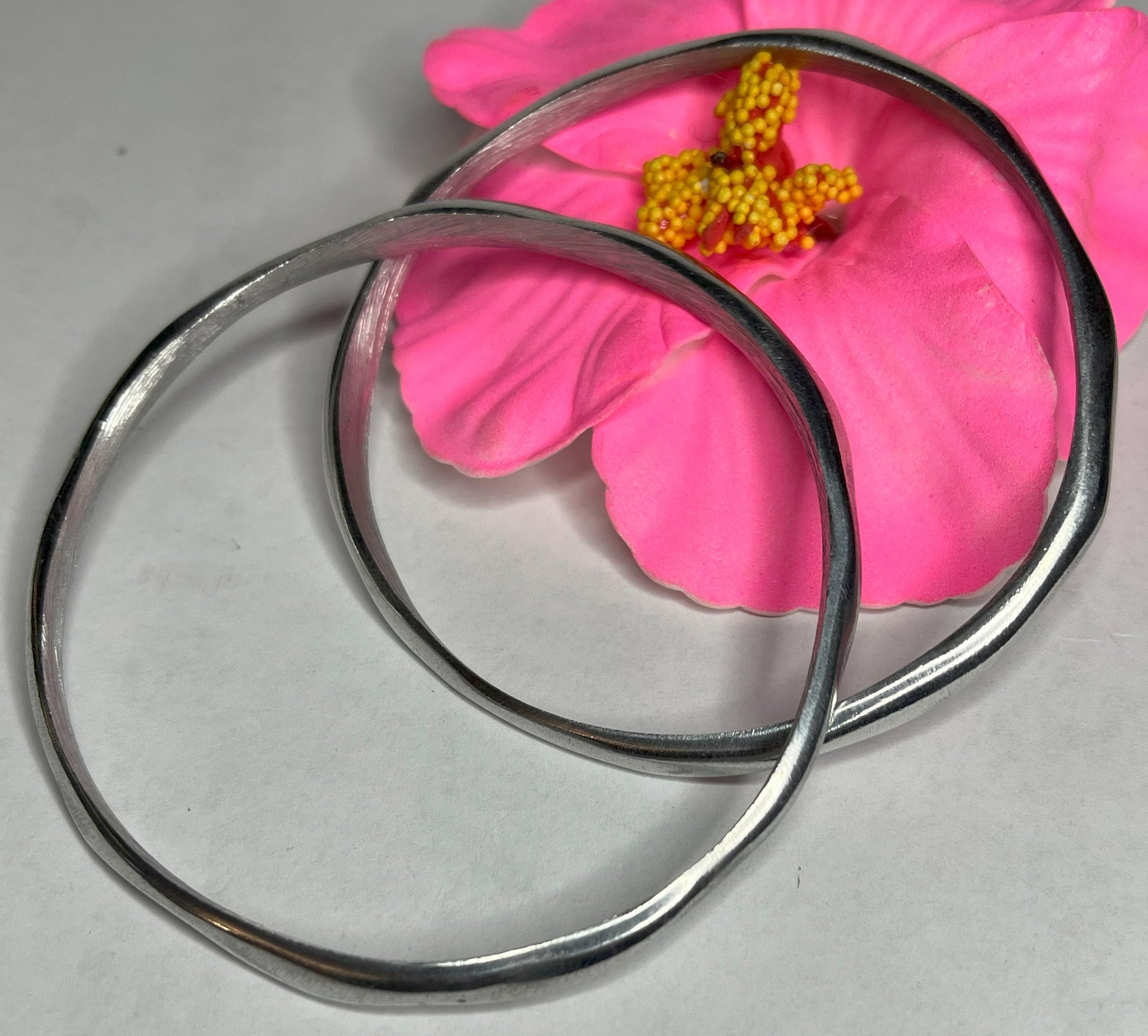 Braccialetto Rinascita in alluminio riciclato fatto a mano, design circolare minimalista con finitura lucida premium, fotografato accanto a un fiore rosa su sfondo neutro