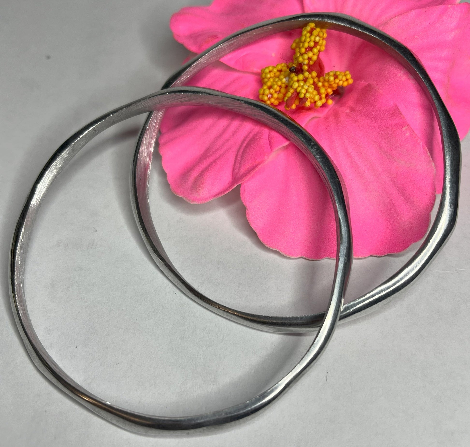 Braccialetto Rinascita in alluminio riciclato fatto a mano, design circolare minimalista con finitura lucida premium, fotografato accanto a un fiore rosa su sfondo neutro