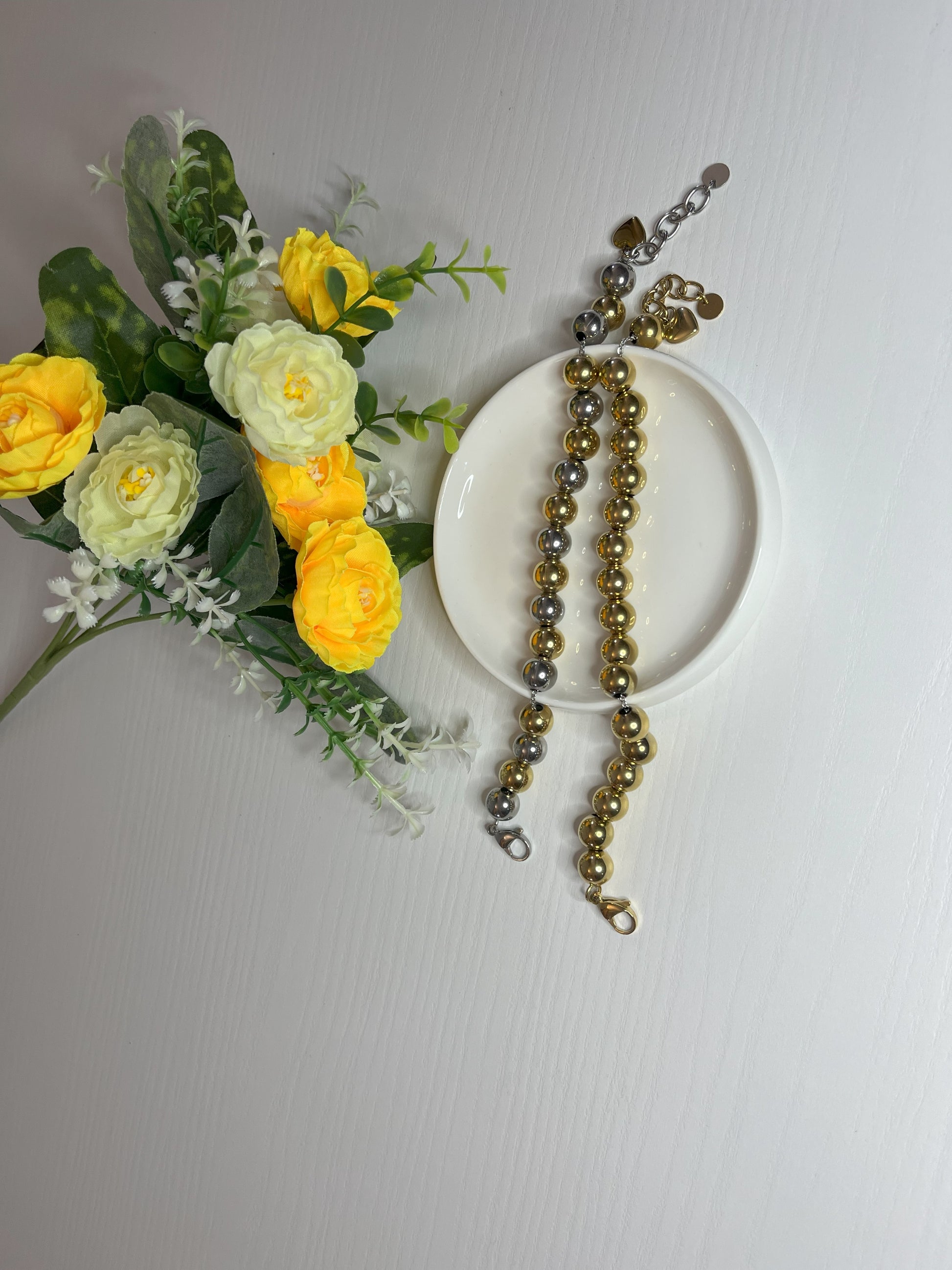 Eleganti braccialetti con sfere dorate e argentate lucide su fiori gialli e foglie verdi