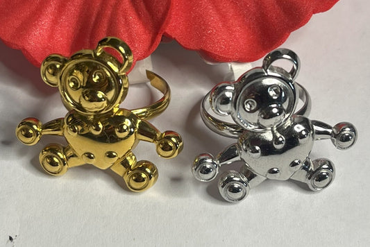 Due anelli con ciondoli a forma di orsetto, uno placcato oro e uno placcato argento, su sfondo bianco con fiore rosso decorativo