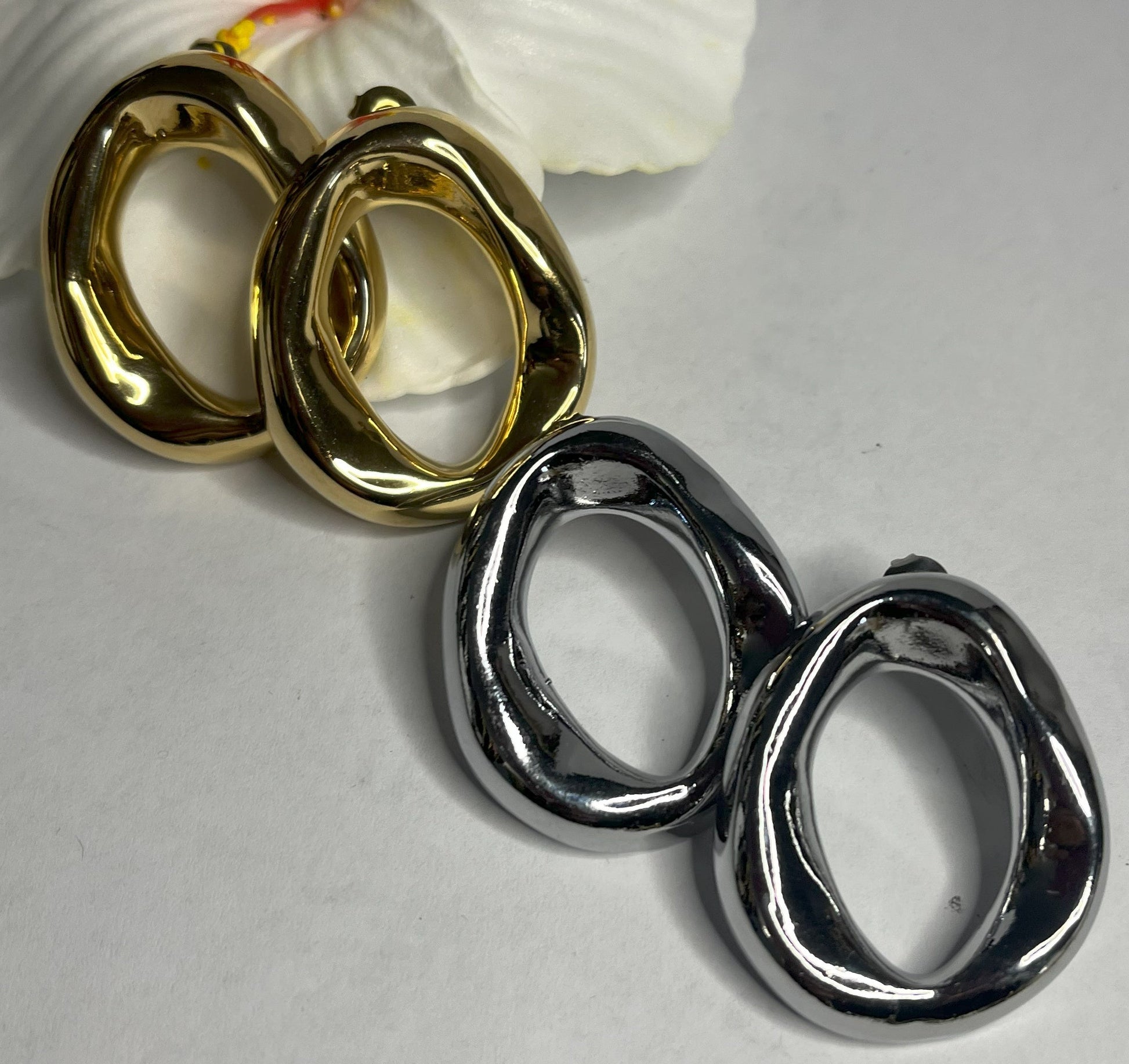 Orecchini Wave premium in due varianti - placcati oro e argento con design organico fluido a forma di onda, fotografati accanto a fiore bianco