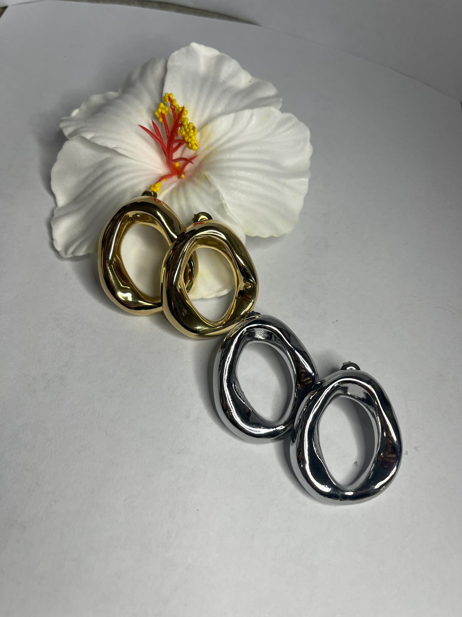 Orecchini Wave premium in due varianti - placcati oro e argento con design organico fluido a forma di onda, fotografati accanto a fiore bianco