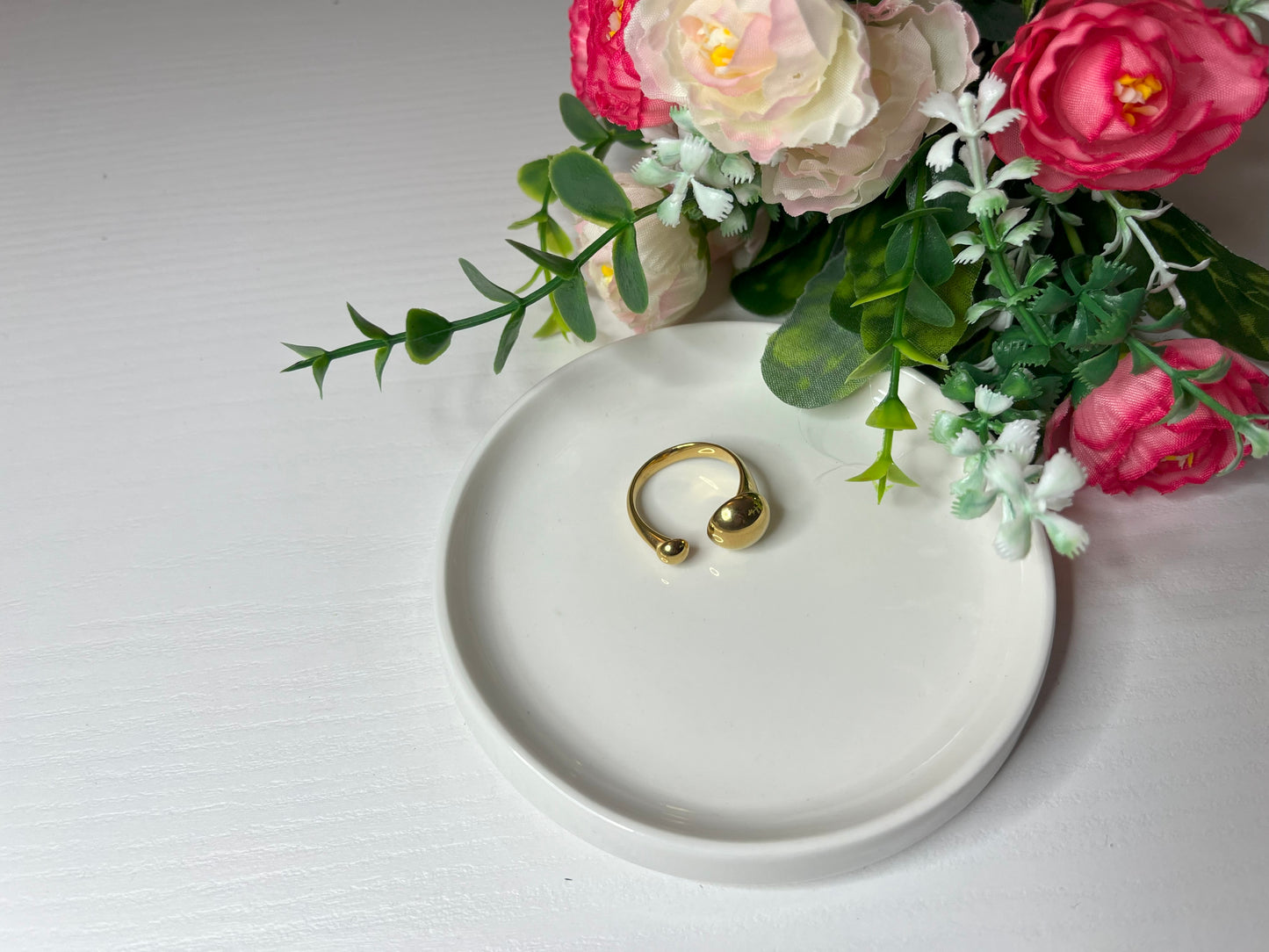 Due anelli minimalisti, uno dorato e uno argentato, posizionati su un piatto bianco accanto a un bouquet di fiori rosa e bianchi