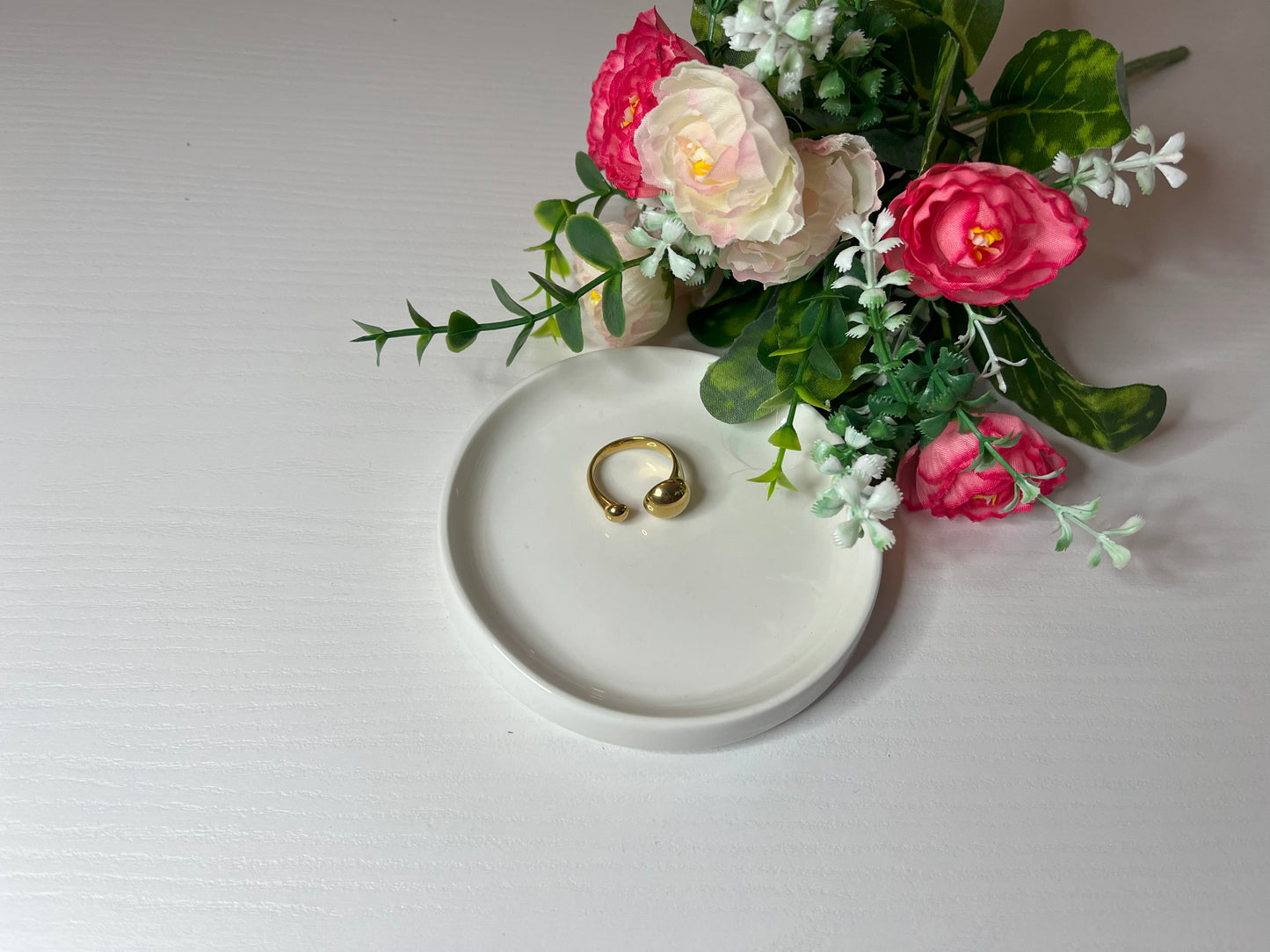 Due anelli minimalisti, uno dorato e uno argentato, posizionati su un piatto bianco accanto a un bouquet di fiori rosa e bianchi