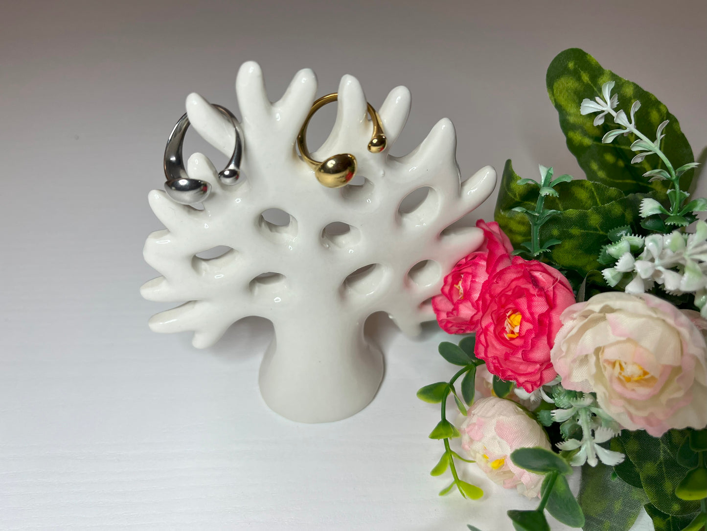 Due anelli minimalisti, uno dorato e uno argentato, posizionati su un piatto bianco accanto a un bouquet di fiori rosa e bianchi