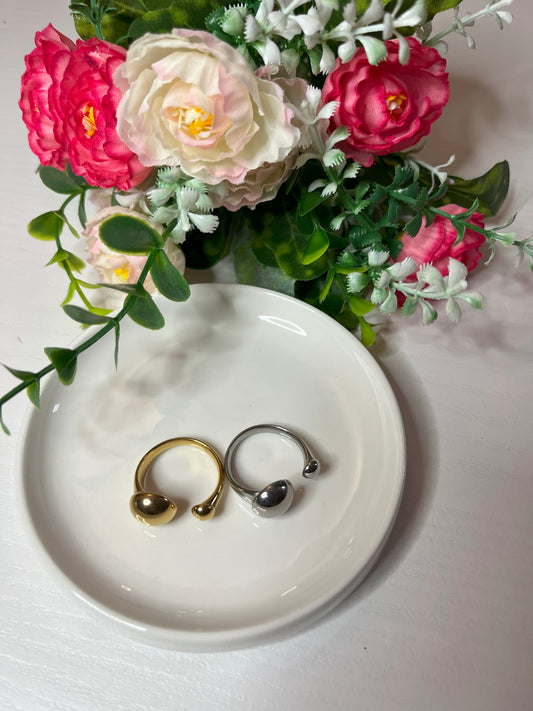 Due anelli minimalisti, uno dorato e uno argentato, posizionati su un piatto bianco accanto a un bouquet di fiori rosa e bianchi