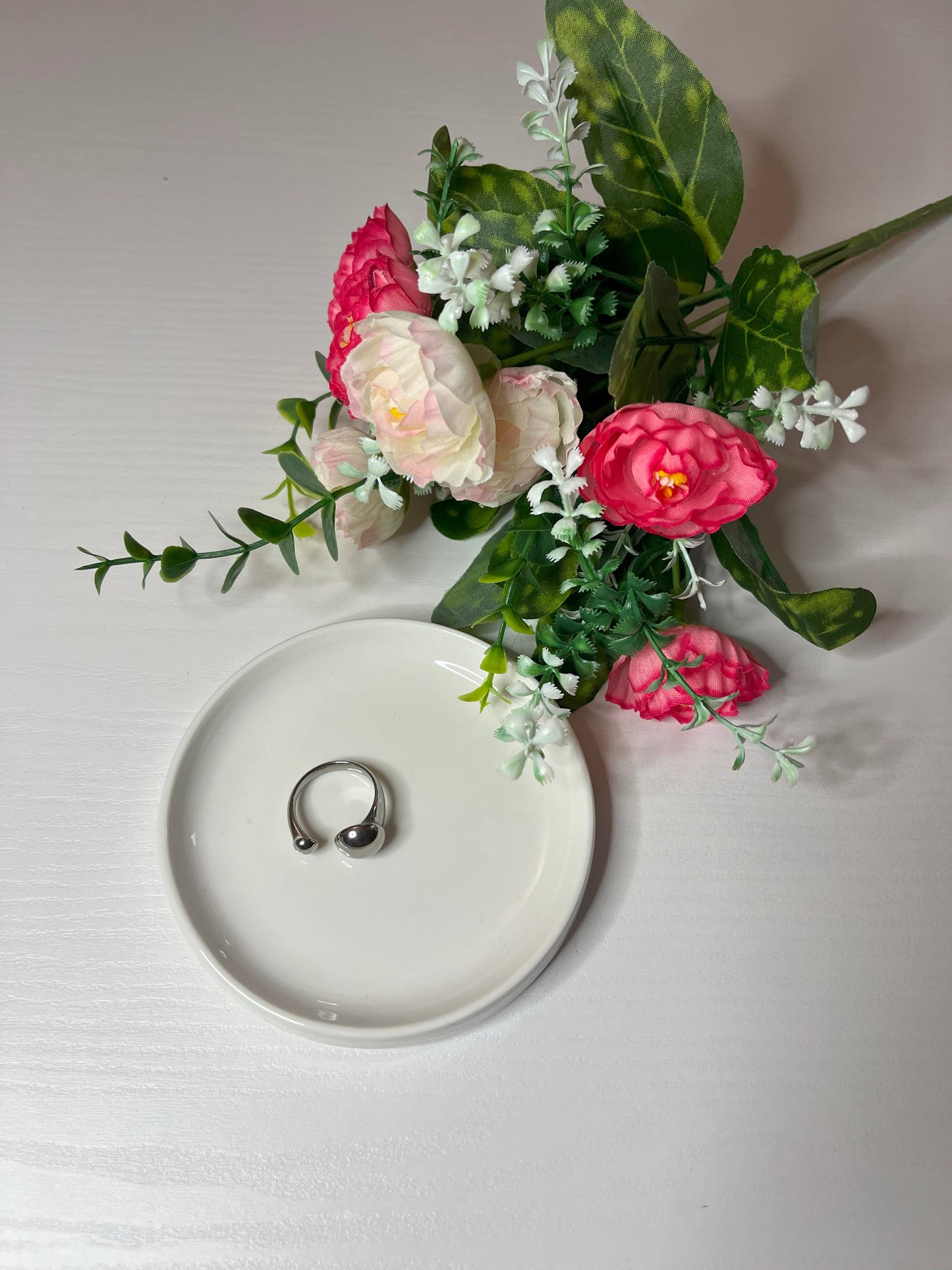 Due anelli minimalisti, uno dorato e uno argentato, posizionati su un piatto bianco accanto a un bouquet di fiori rosa e bianchi