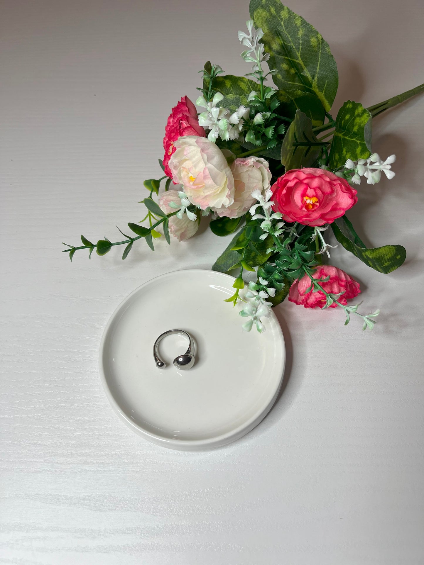 Due anelli minimalisti, uno dorato e uno argentato, posizionati su un piatto bianco accanto a un bouquet di fiori rosa e bianchi