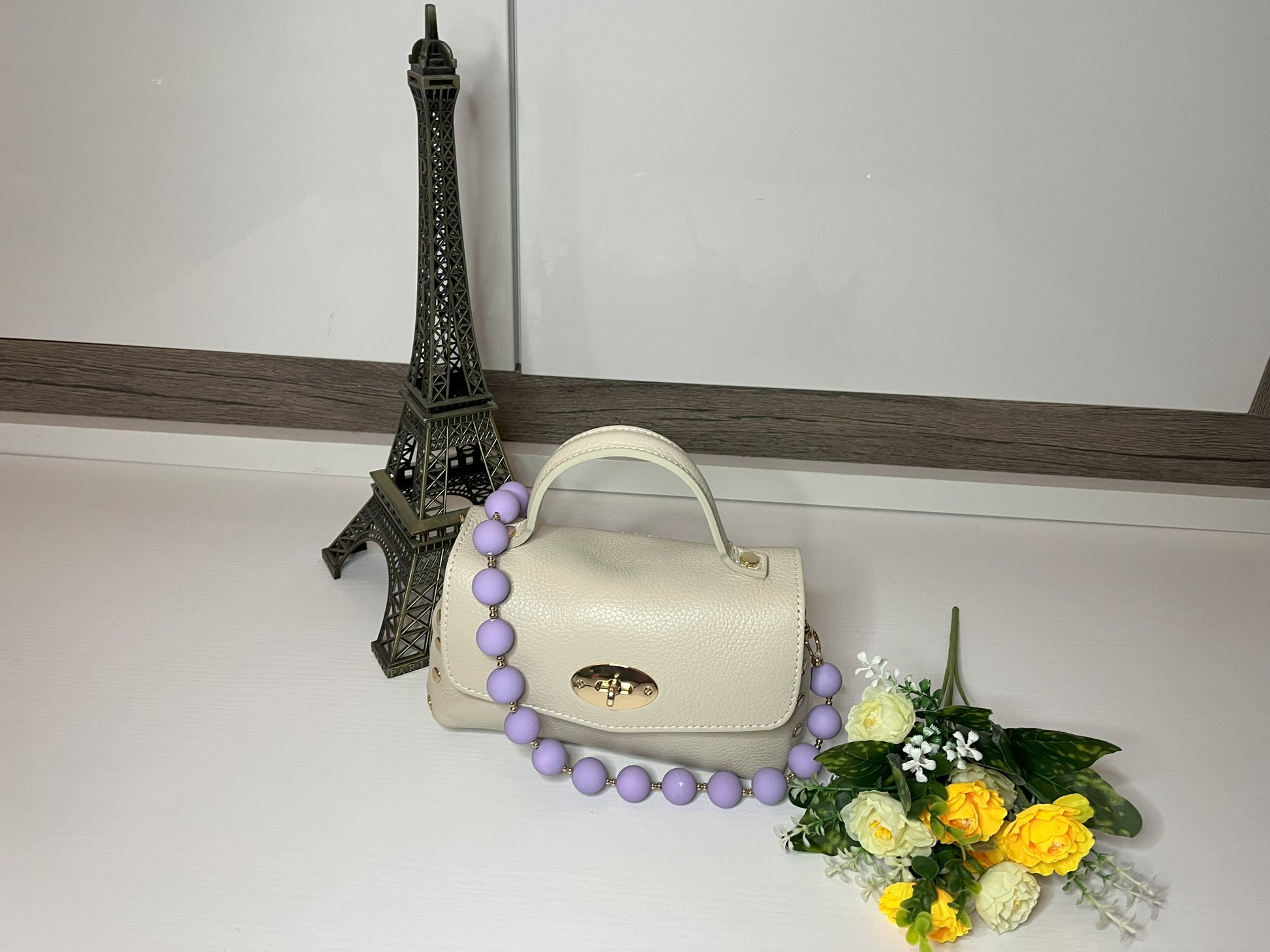 Mini-Parisienne borsa beige compatta con collana di perle viola coordinata, accanto a Torre Eiffel decorativa e fiori gialli
