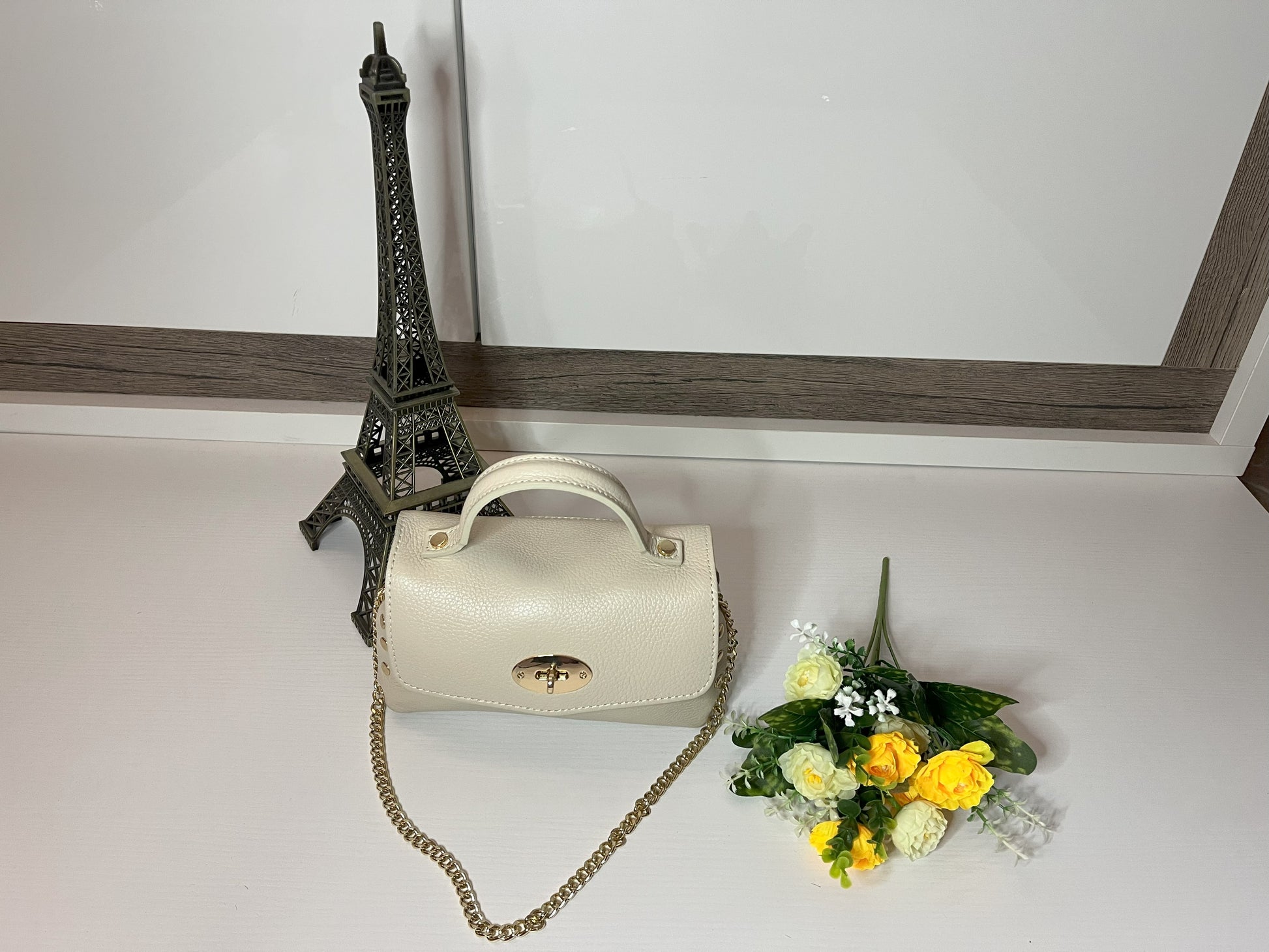 Mini-Parisienne borsa beige compatta con collana di perle viola coordinata, accanto a Torre Eiffel decorativa e fiori gialli