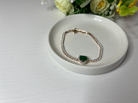 Elegante braccialetto tennis con cristalli brillanti e pietra verde smeraldo centrale, indossato al polso su sfondo tessuto bianco