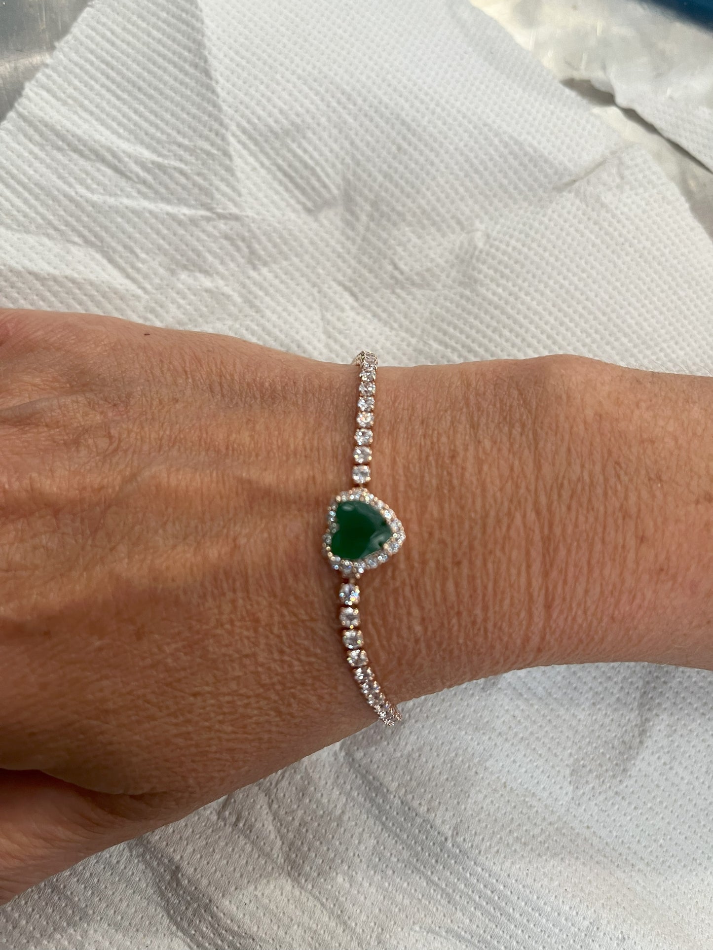 Elegante braccialetto tennis con cristalli brillanti e pietra verde smeraldo centrale, indossato al polso su sfondo tessuto bianco