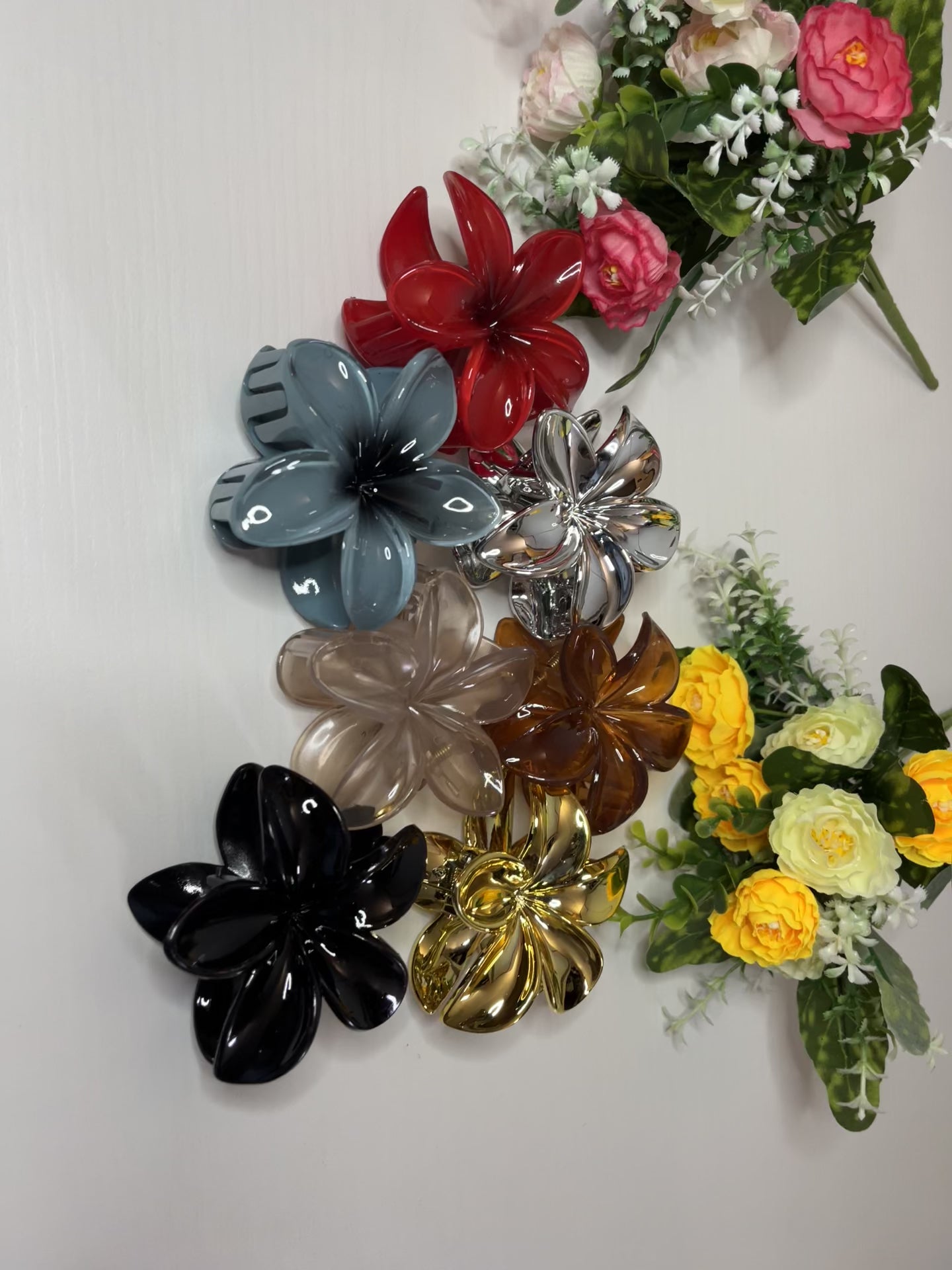 Elegante pinza hawaiana per capelli a forma di fiore con petali lucidi, posizionata accanto a un bouquet di fiori freschi gialli e bianchi