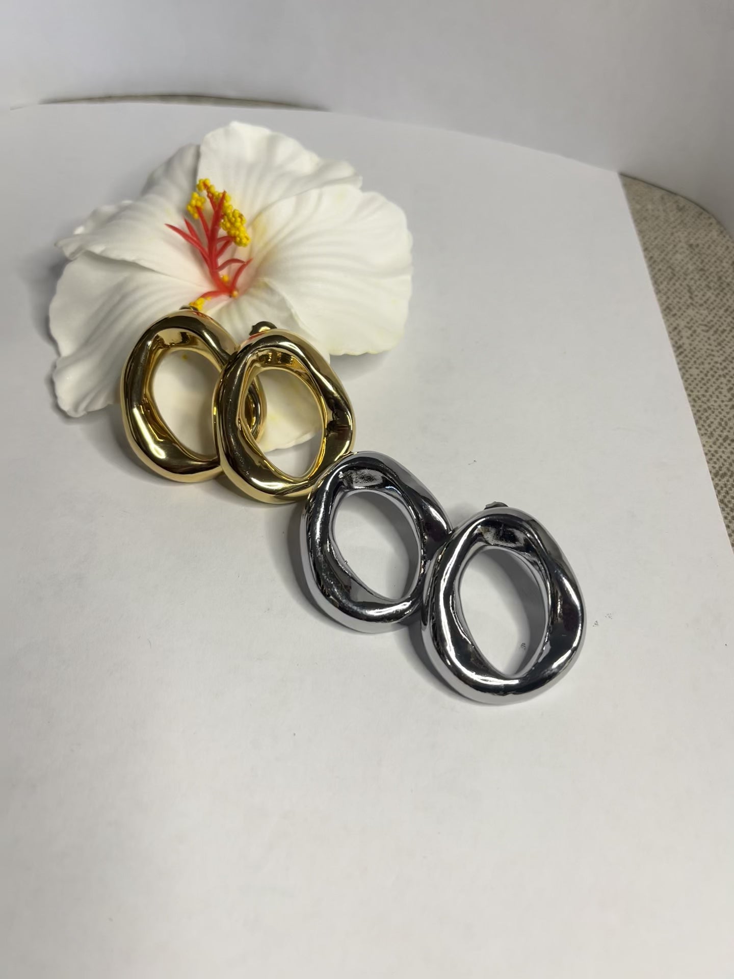 Orecchini Wave premium in due varianti - placcati oro e argento con design organico fluido a forma di onda, fotografati accanto a fiore bianco