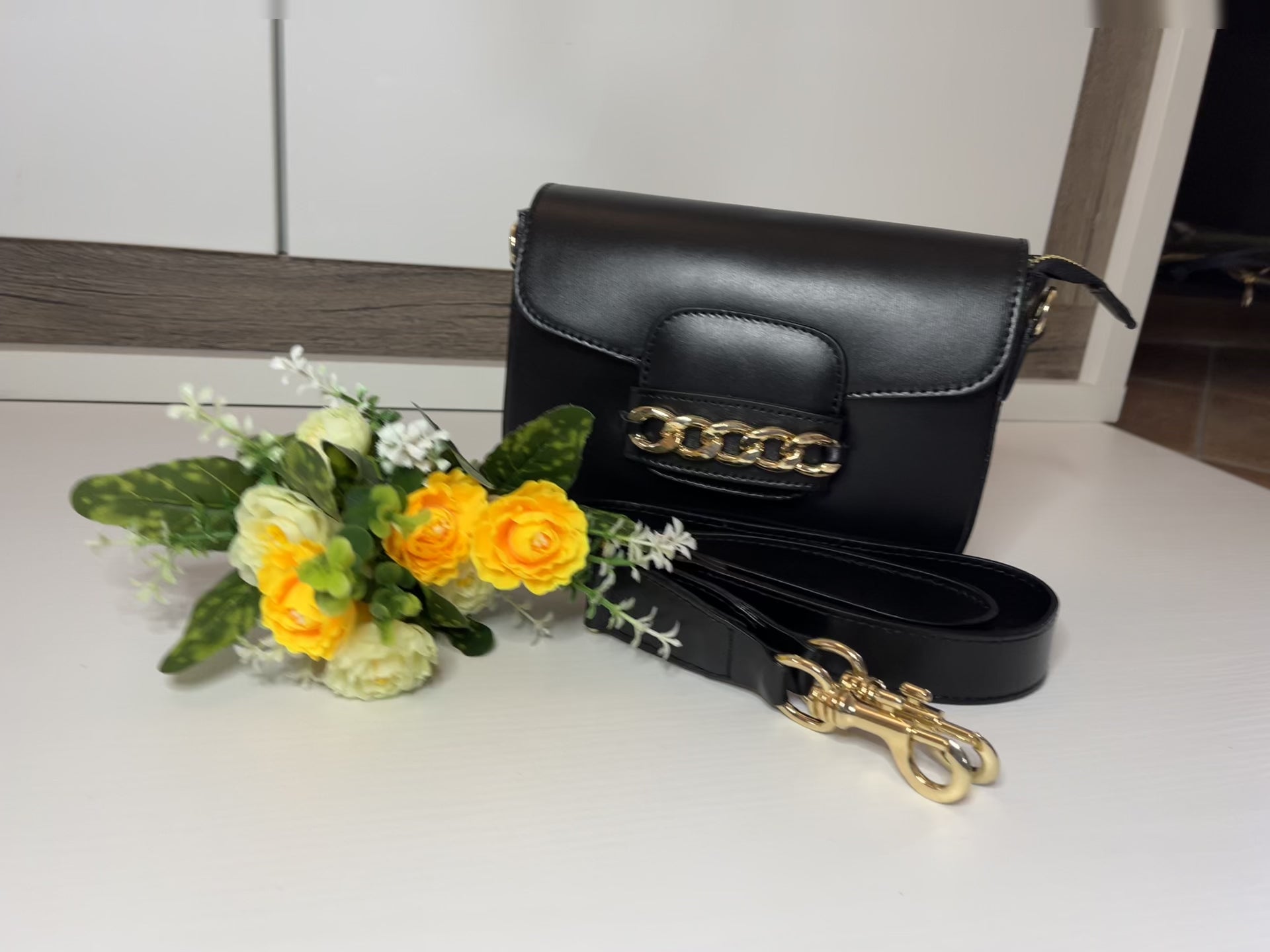 Borsa Toscana Chic in vera pelle nera Made in Italy con catena dorata decorativa e tracolla regolabile, fotografata su superficie bianca con fiori gialli