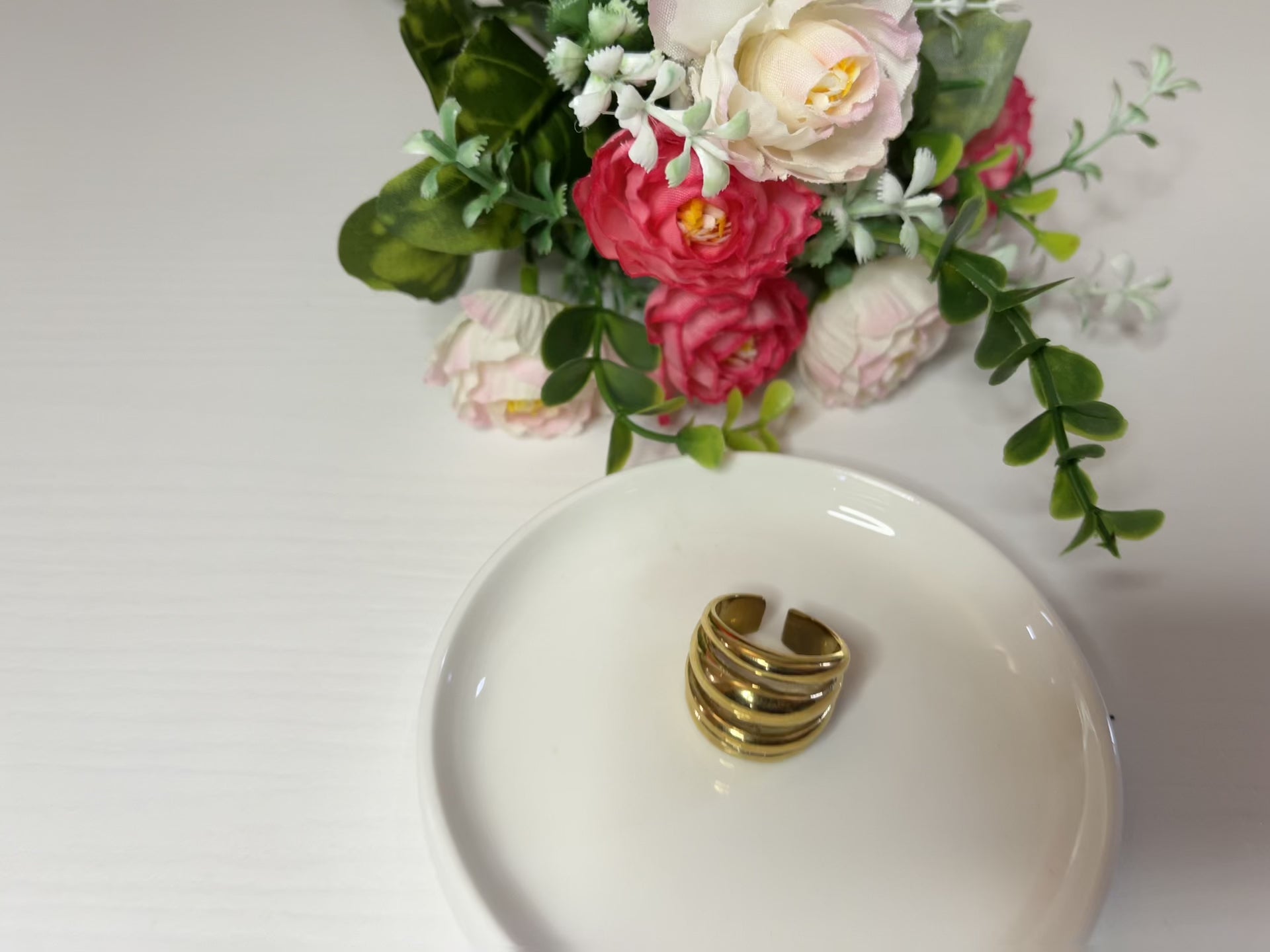 Anello Fascia Tripla placcato oro con tre fasce sovrapposte eleganti, design moderno e minimalista su piattino bianco con fiore rosa e bianco