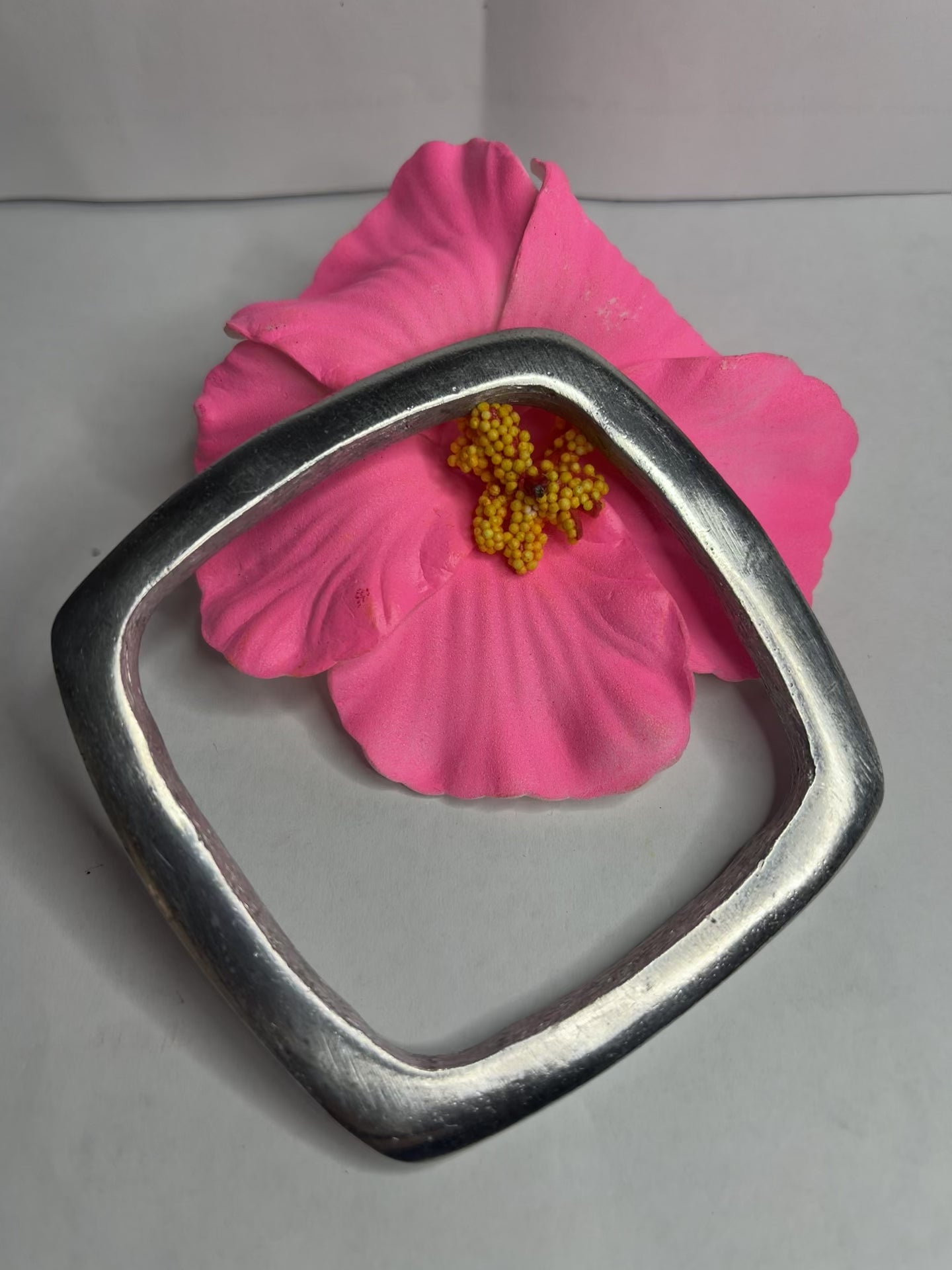Bracciale quadro in alluminio con design geometrico moderno e finitura satinata, fotografato accanto a fiore rosa