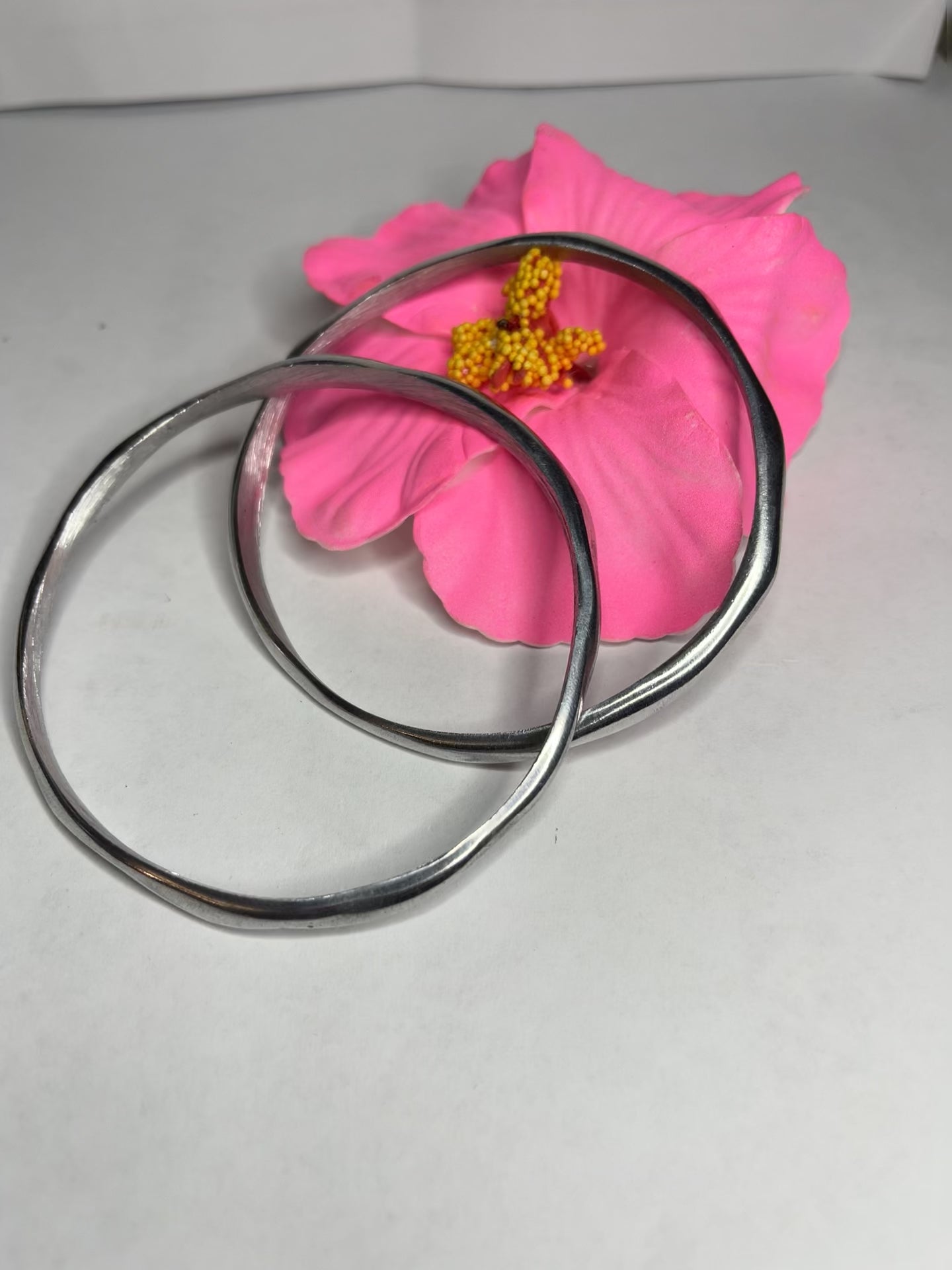 Braccialetto Rinascita in alluminio riciclato fatto a mano, design circolare minimalista con finitura lucida premium, fotografato accanto a un fiore rosa su sfondo neutro