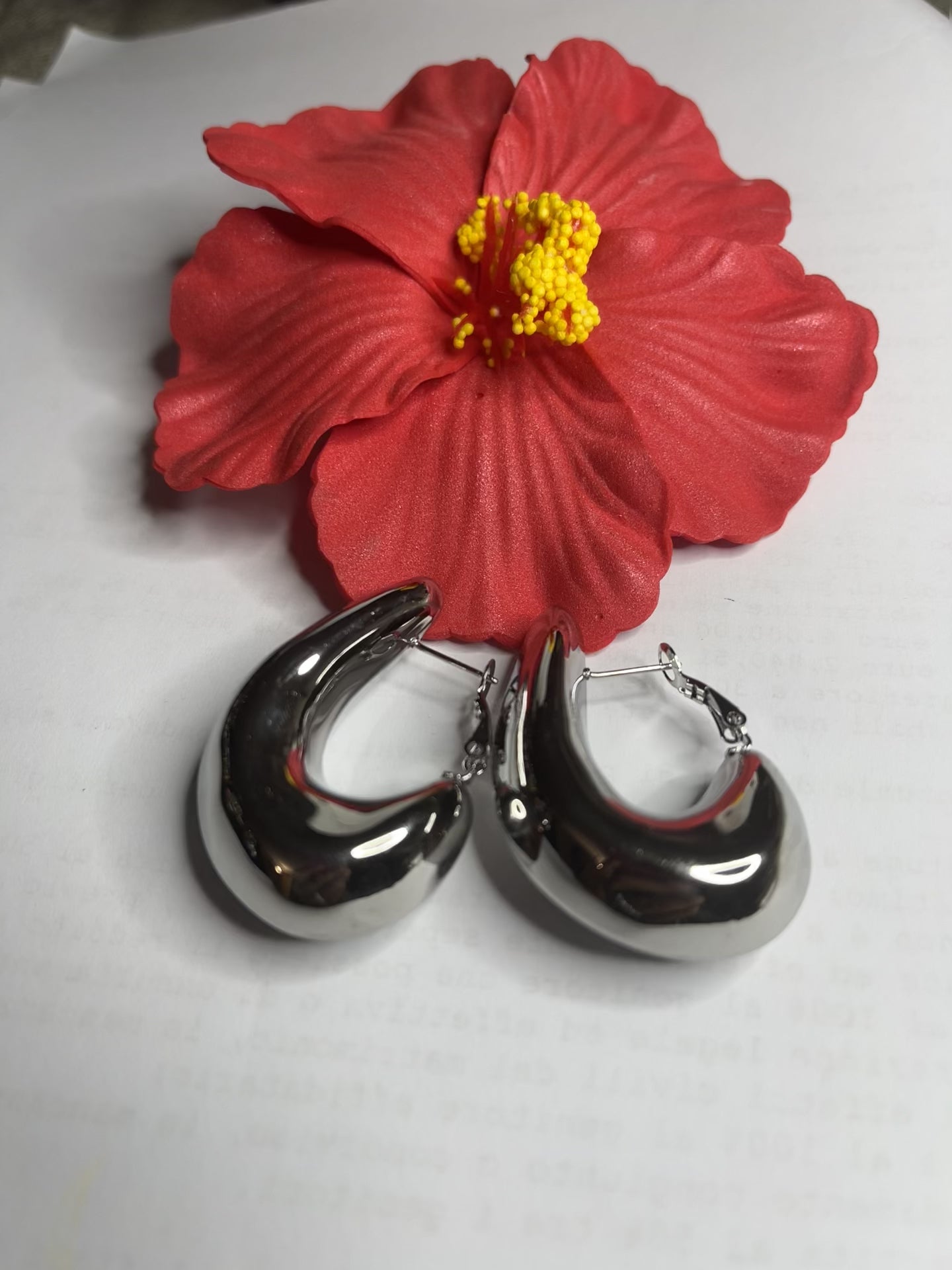 Orecchini Della Schiava a cerchio in acciaio premium placcato argento con superficie liscia e brillante, chiusura a scatto sicura, fotografati su superficie bianca e fiore rosso