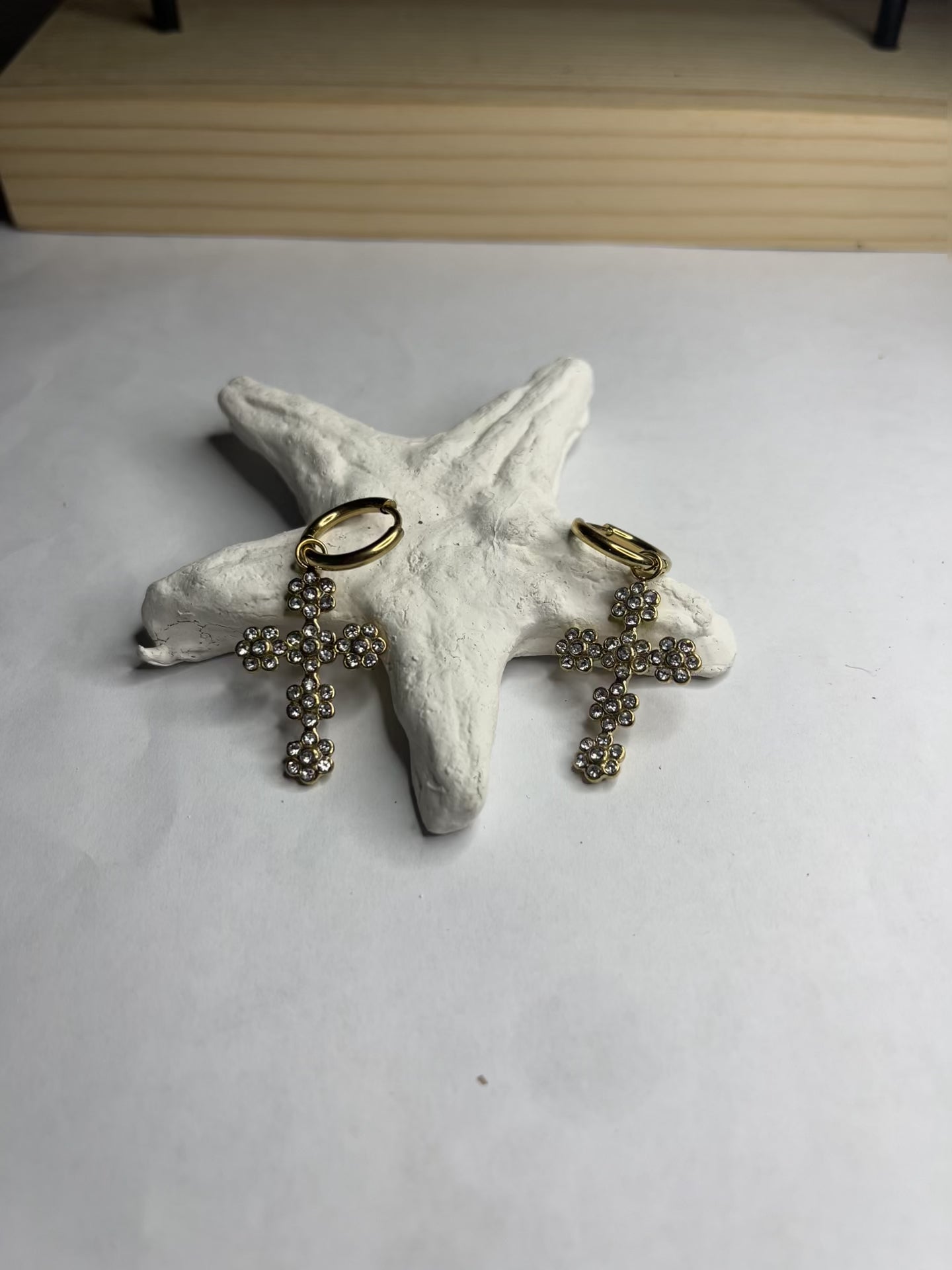 Orecchini pendenti con ciondoli a croce decorati con cristalli diamantati e ganci dorati, appoggiati su stella marina bianca