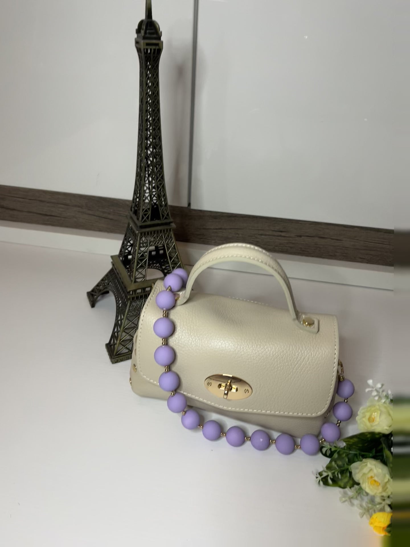 Mini-Parisienne borsa beige compatta con collana di perle viola coordinata, accanto a Torre Eiffel decorativa e fiori gialli