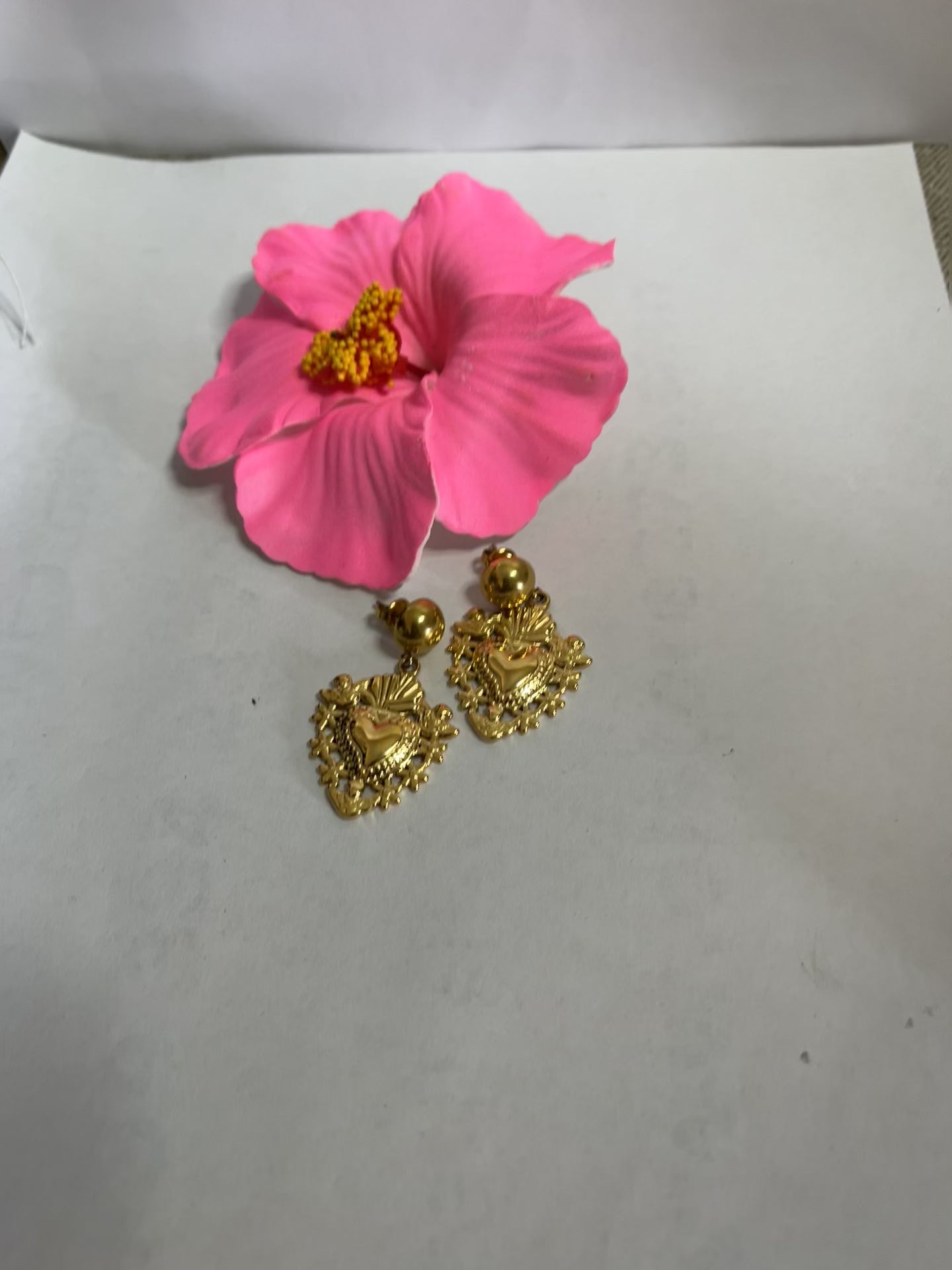 Orecchini dorati a forma di sacro cuore con dettagli intarsiati e lavorazione artigianale su sfondo con fiore rosa