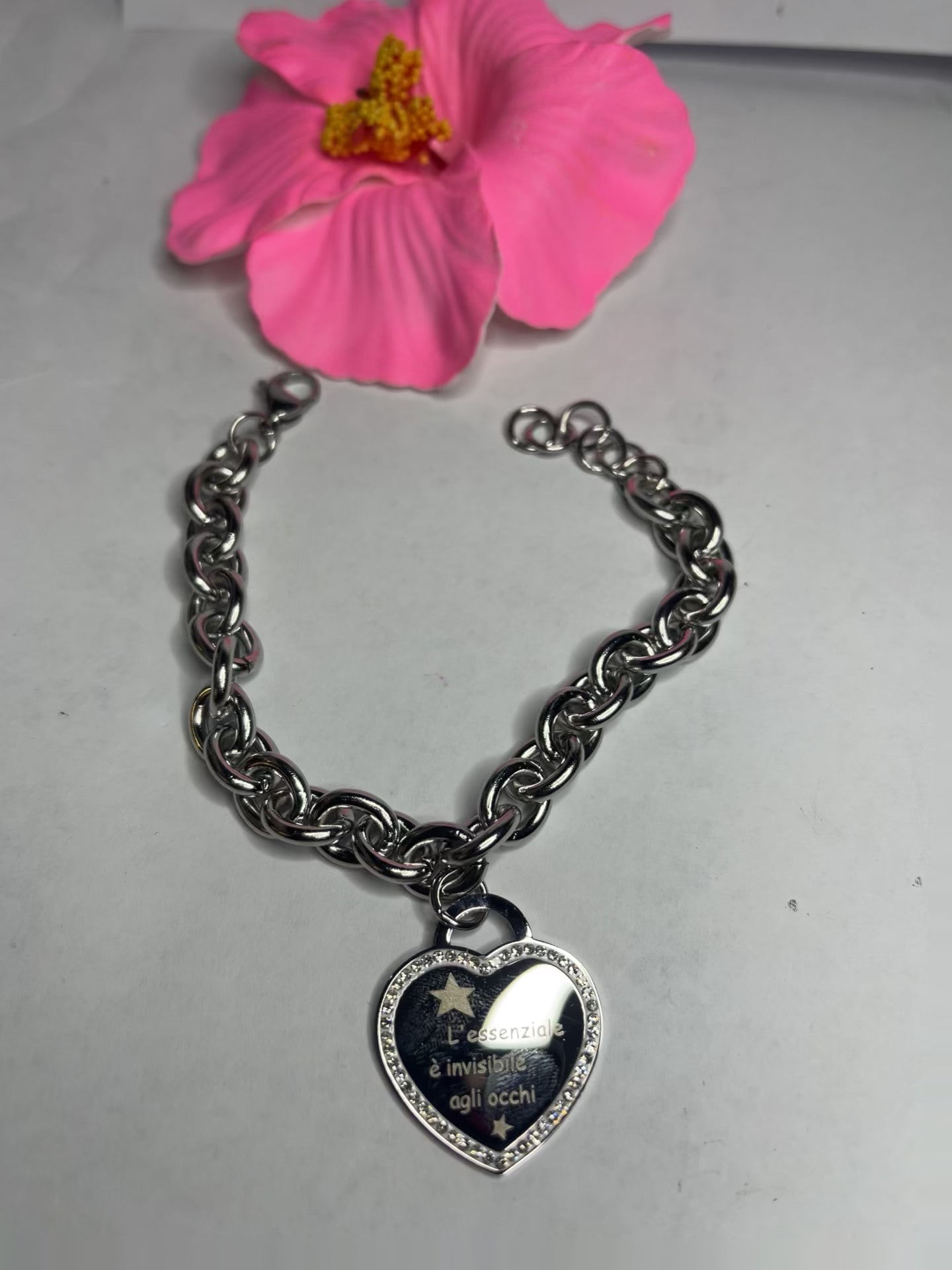 Braccialetto chunky argentato con pendente a cuore nero contenente la scritta "L'essenziale è invisibile agli occhi" su sfondo grigio con petali rosa