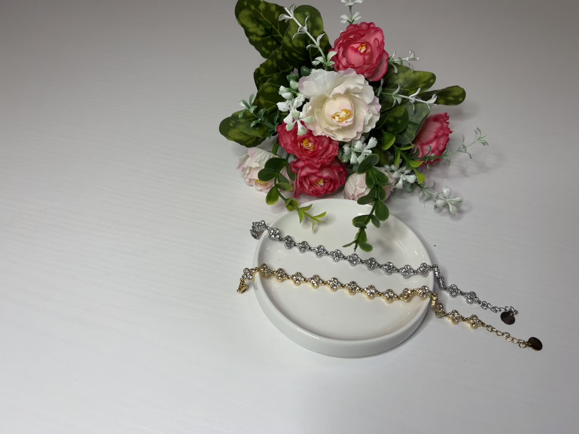Braccialetti tennis, uno placcato oro, uno placcato argento con quadrifogli portafortuna e cristalli scintillanti su sfondo bianco con foglie verdi