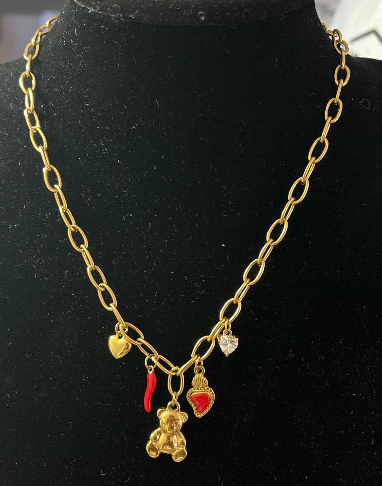 Rita - Collana placcata oro con catena e ciondoli portafortuna tra cui orsetto, cornetto rosso e cuori, esposta su busto nero