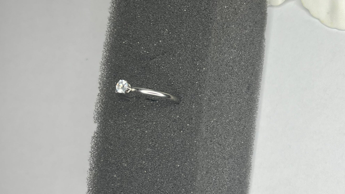 Anello solitario in acciaio placcato argento con pietra brillante centrale, montatura classica elegante su superficie grigia con conchiglia decorativa