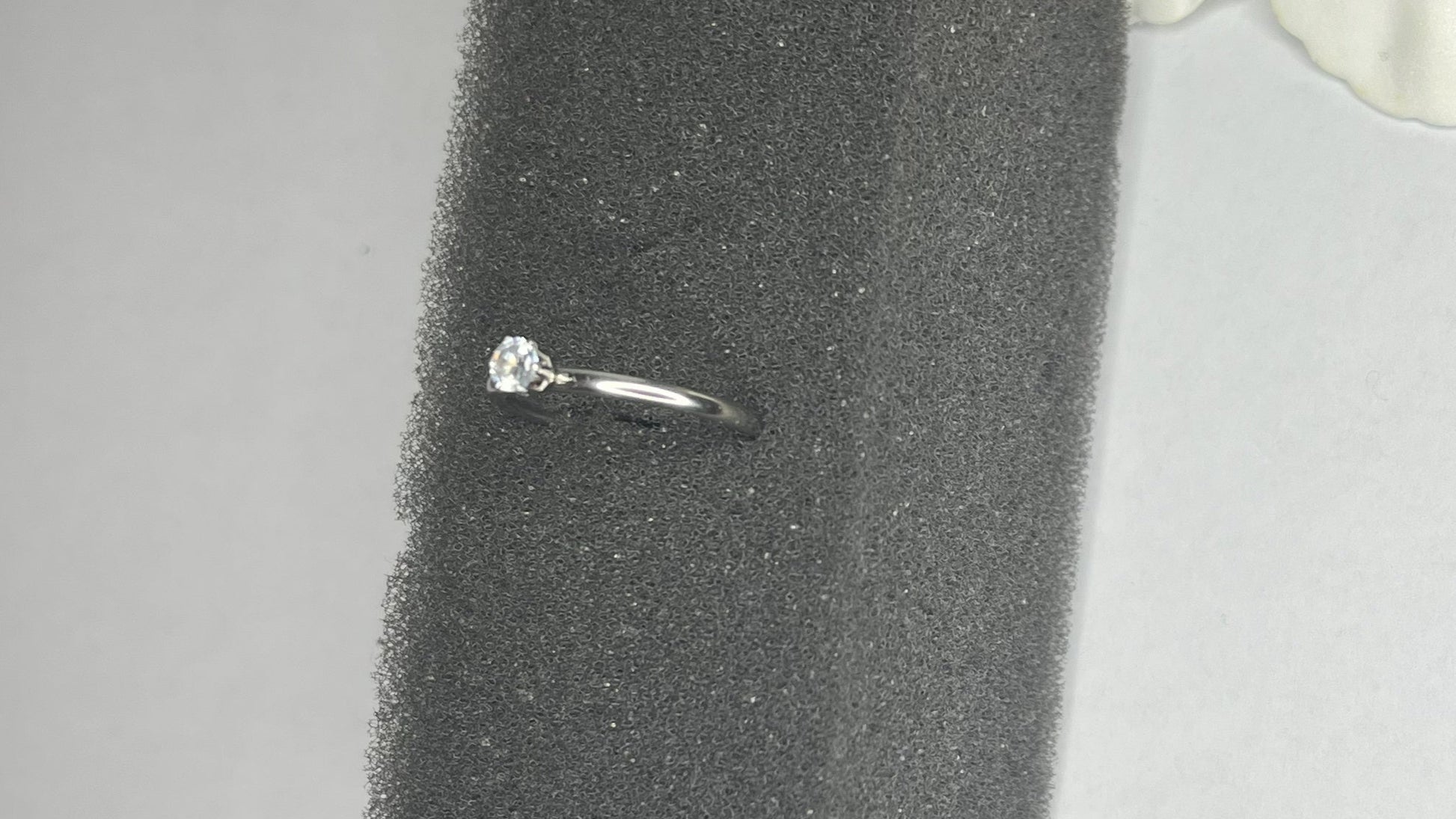 Anello solitario in acciaio placcato argento con pietra brillante centrale, montatura classica elegante su superficie grigia con conchiglia decorativa